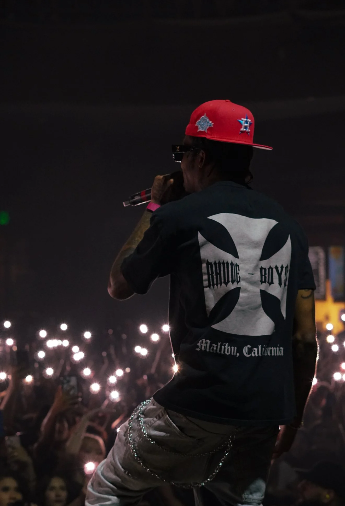 A male performer on stage wearing a black T-shirt with 'Bauace Boyz Malibu, California' written on it, a red cap, sunglasses, and holding a microphone, during a concert with an audience holding up lights in the background.