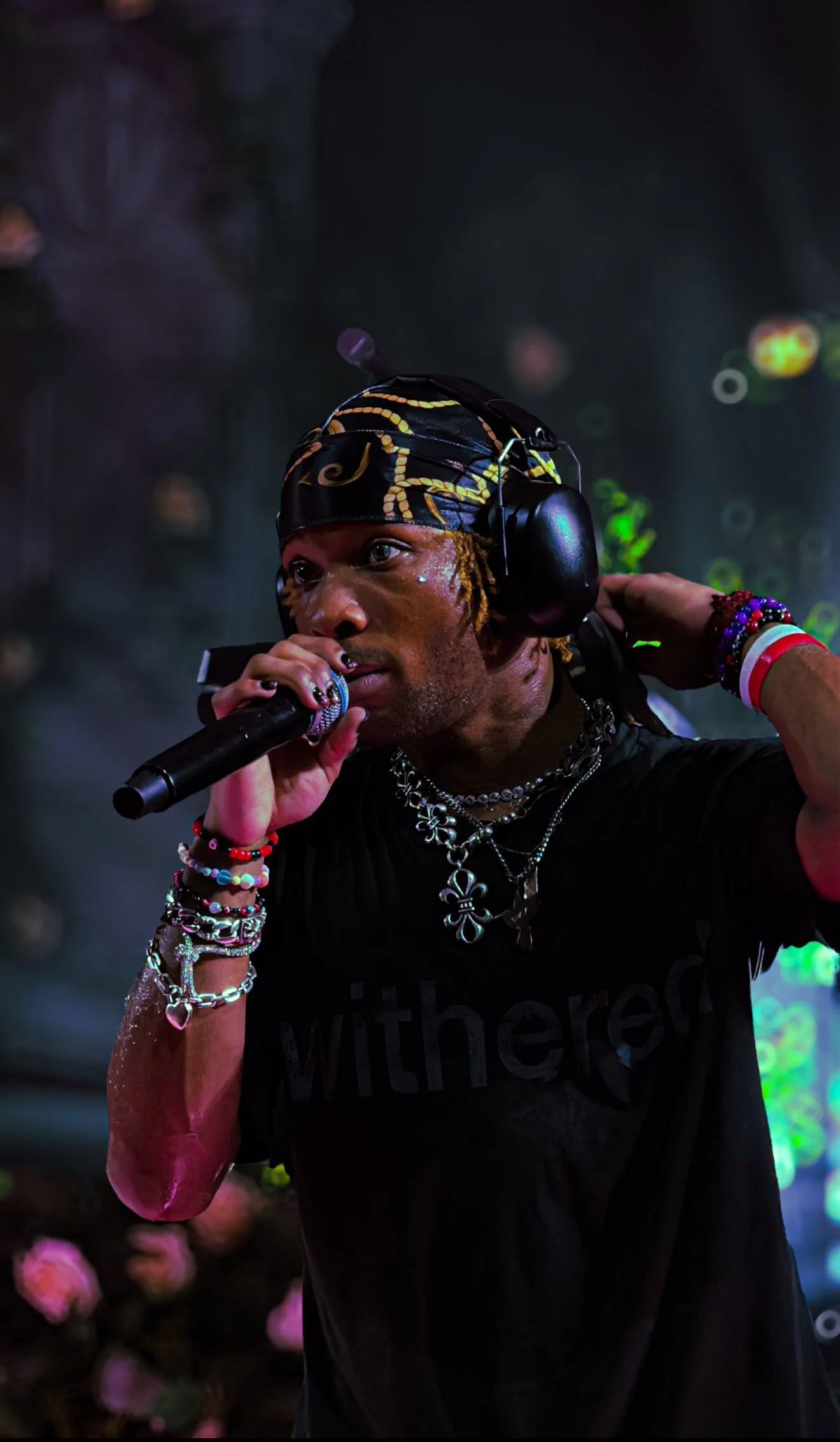 A person wearing a black T-shirt, decorated with jewelry and bracelets, is speaking into a microphone, wearing large headphones, and a colorful headscarf with yellow patterns, while performing on stage with blurry colorful lights and floral decoratio