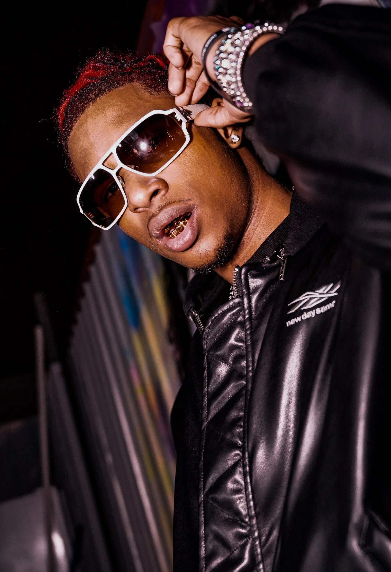 A young man wearing large sunglasses, gold grills on his teeth, a black leather jacket, and jewelry, posing in front of a colorful graffiti wall.