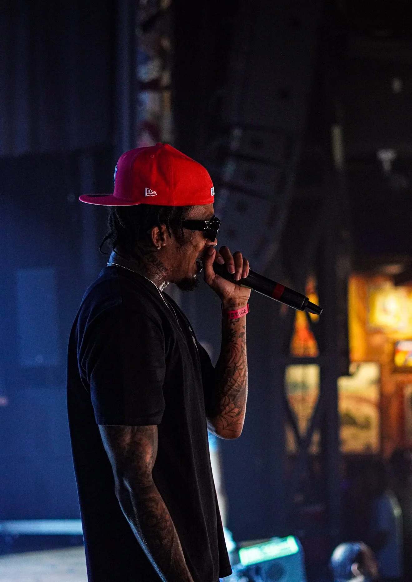 A male performer with tattoos on his arm, wearing sunglasses and a red cap, holding a microphone on stage in a dimly lit venue.