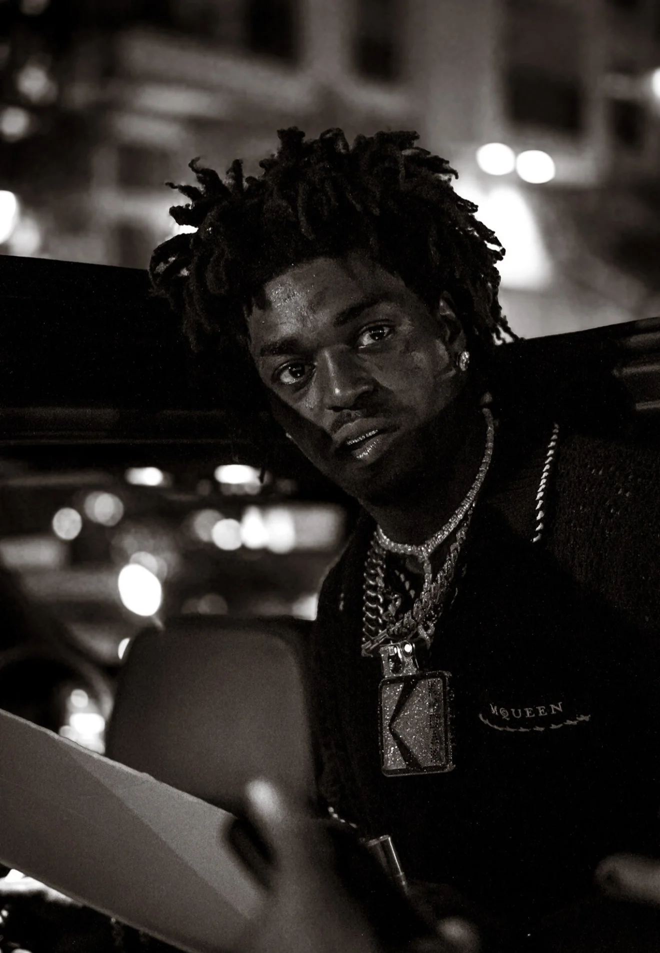 A young man with dreadlocks and multiple necklaces, including a large pendant, looking directly at the camera in a dark, urban setting at night.