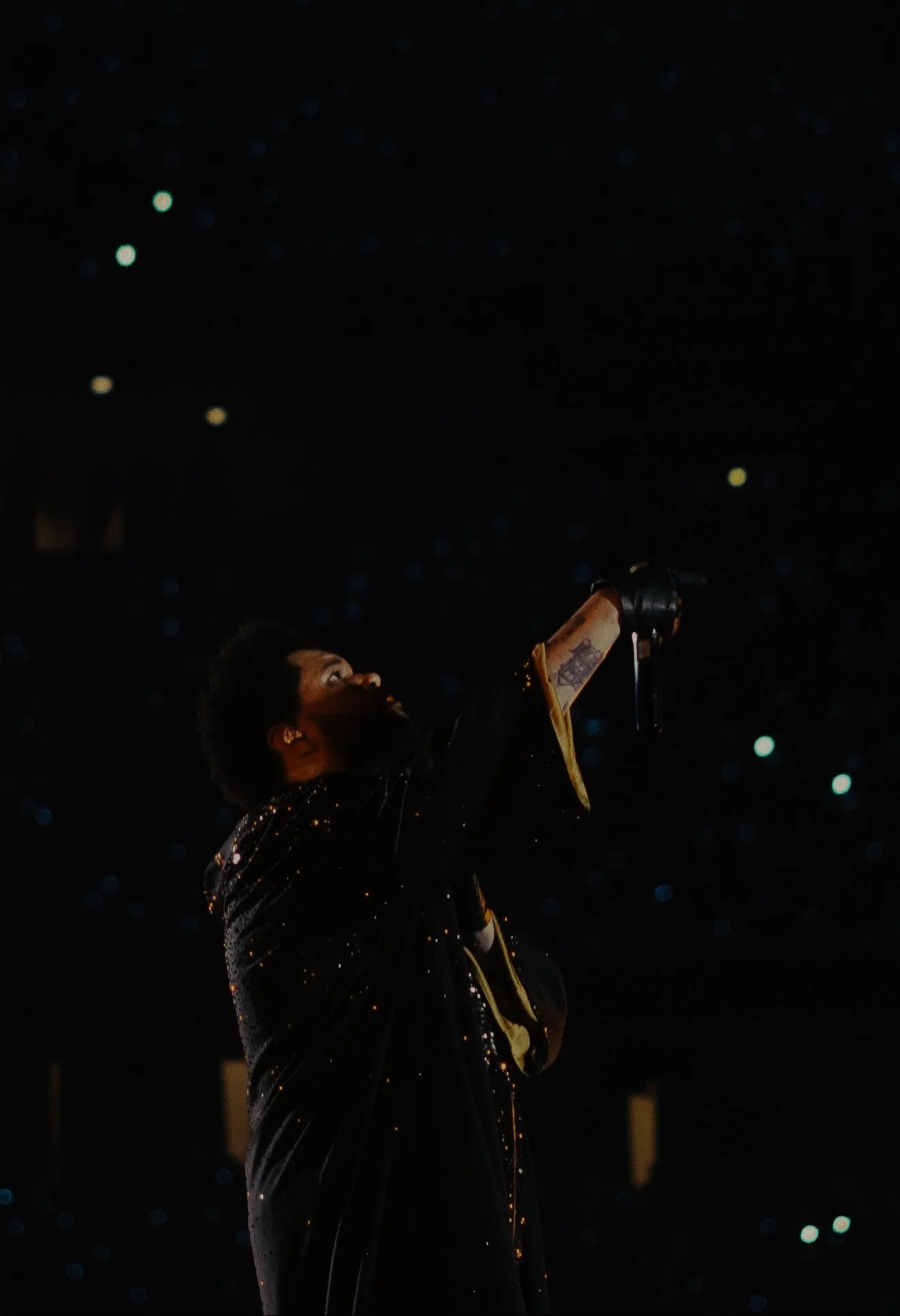 A performer on stage in a black and gold sequined jacket, holding a microphone, with a dark background and small bright lights.