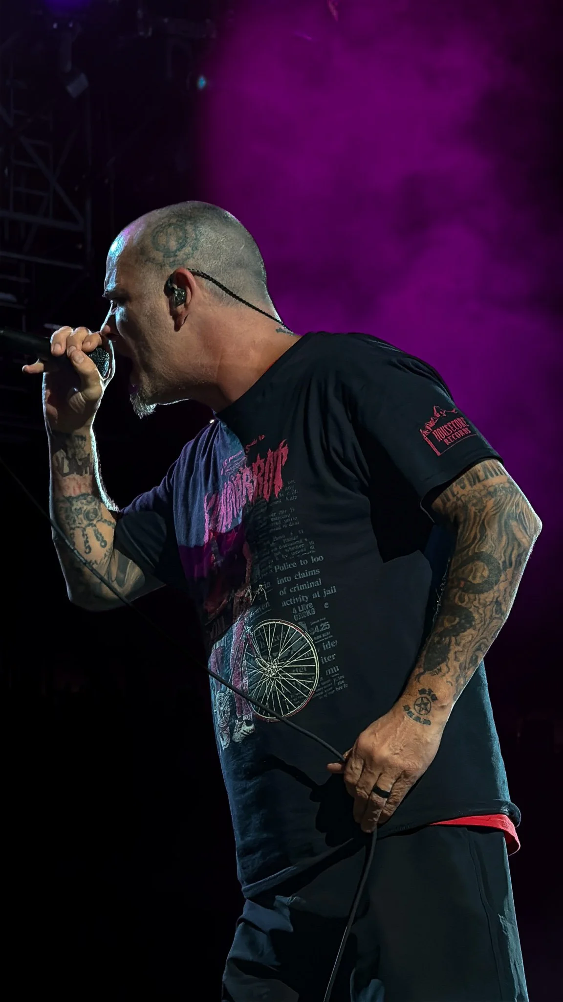 A tattooed male singer with a shaved head singing into a microphone on stage, with purple stage lights in the background.