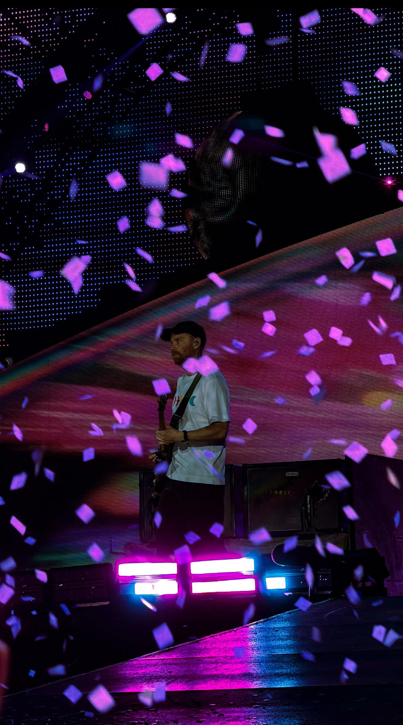 A man playing a guitar on stage amidst falling purple confetti, with vibrant LED lights and digital backgrounds creating a colorful concert atmosphere.