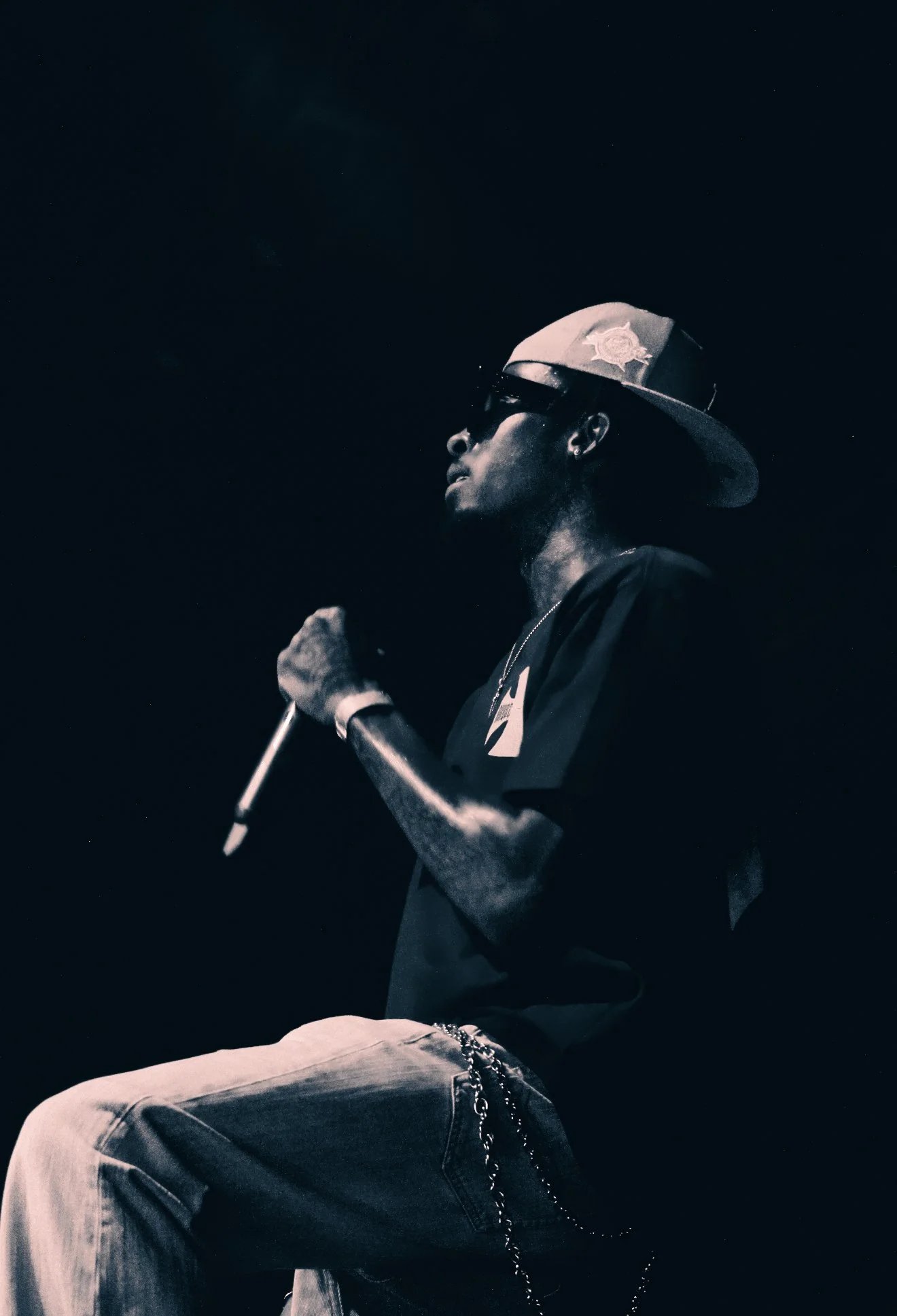 A man wearing sunglasses and a cap with a police badge, holding a microphone, sitting against a dark background.