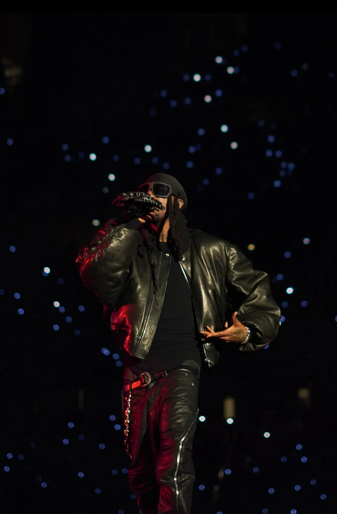 A male performer wearing a black leather jacket, black pants, sunglasses, and a hat, singing into a microphone against a starry background.