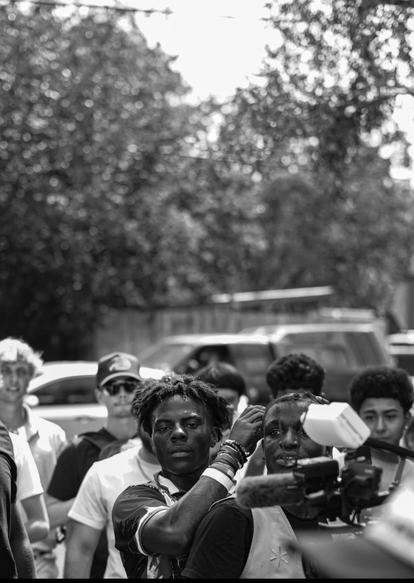 A black and white photo of a diverse group of young people outdoors, some wearing sunglasses and bracelets, with a camera and microphone in front of them; trees and cars are in the background.