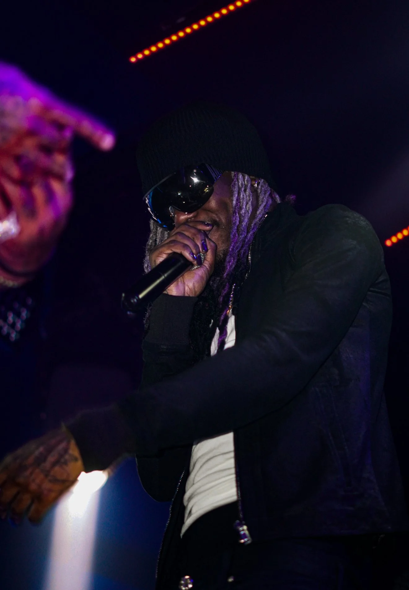 A person with purple dreadlocks, wearing a black beanie, sunglasses, and a black jacket, holding a microphone and performing on stage with purple and orange lights overhead.
