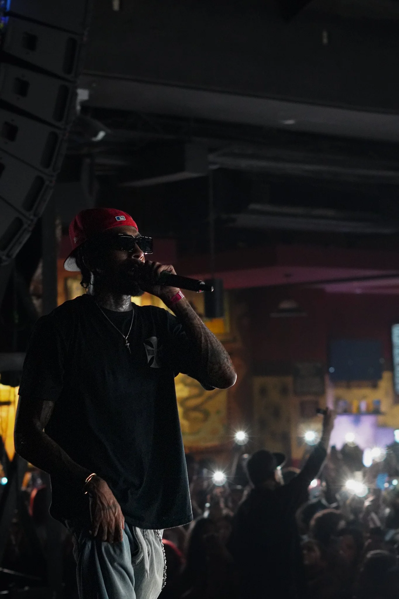 Musician wearing sunglasses and a red baseball cap performing on stage holding a microphone, with an audience in the background holding up phone lights.