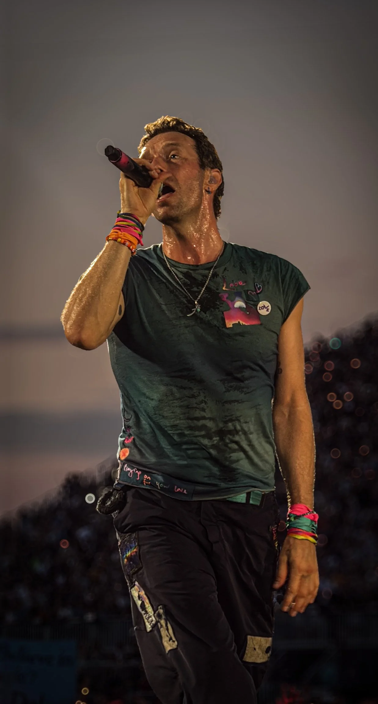 Male performer singing into microphone on stage, sweat visible, wearing a black t-shirt adorned with colorful patches and pins, black pants with patches, multiple colorful wristbands, and jewelry, during a concert at dusk or night.
