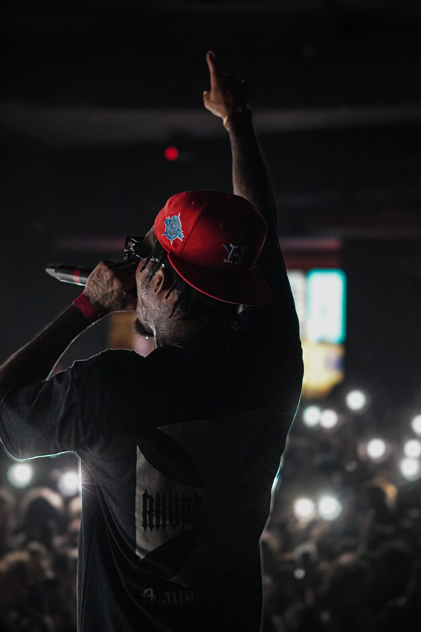 A performer on stage wearing a red cap with police badge emblem, holding a microphone, gesturing upward with one arm, in front of a large audience with many audience members holding up lights.