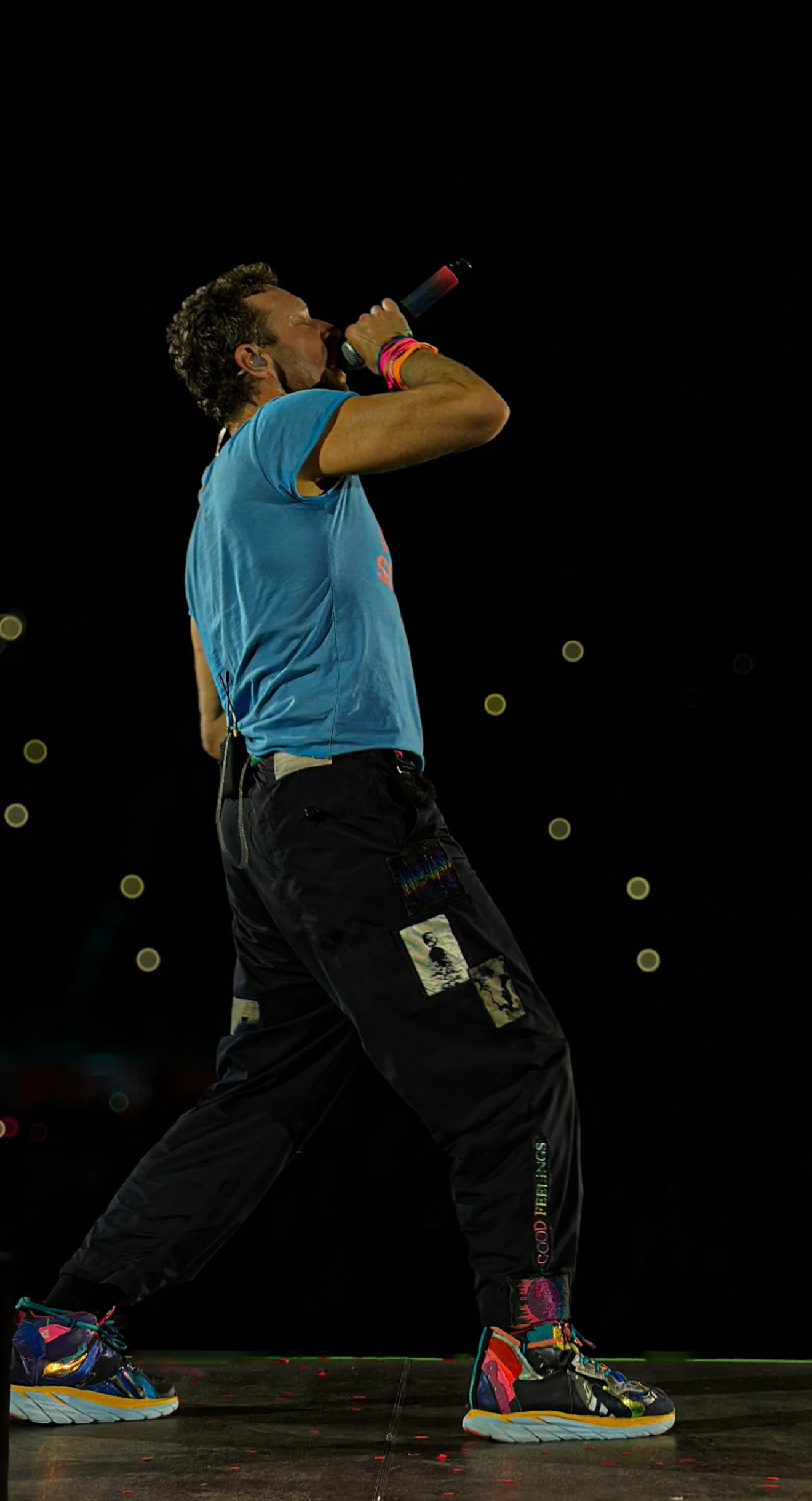 Performer singing into a microphone on stage at night, wearing a blue t-shirt, black pants with patches, and colorful sneakers.