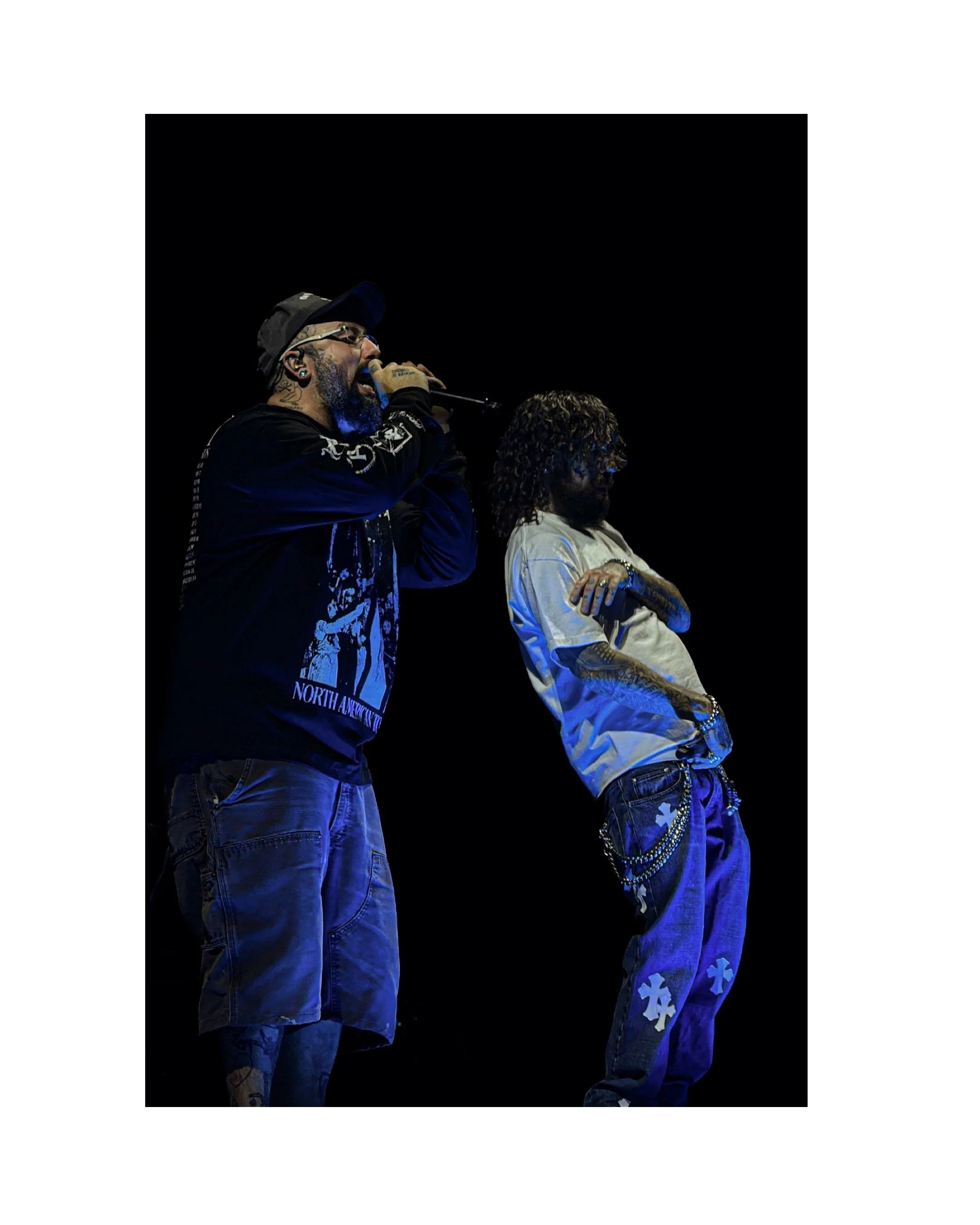 Two male performers on stage, one singing into a microphone with glasses and a cap, the other with long hair and tattoos, standing with crossed arms against a dark background.
