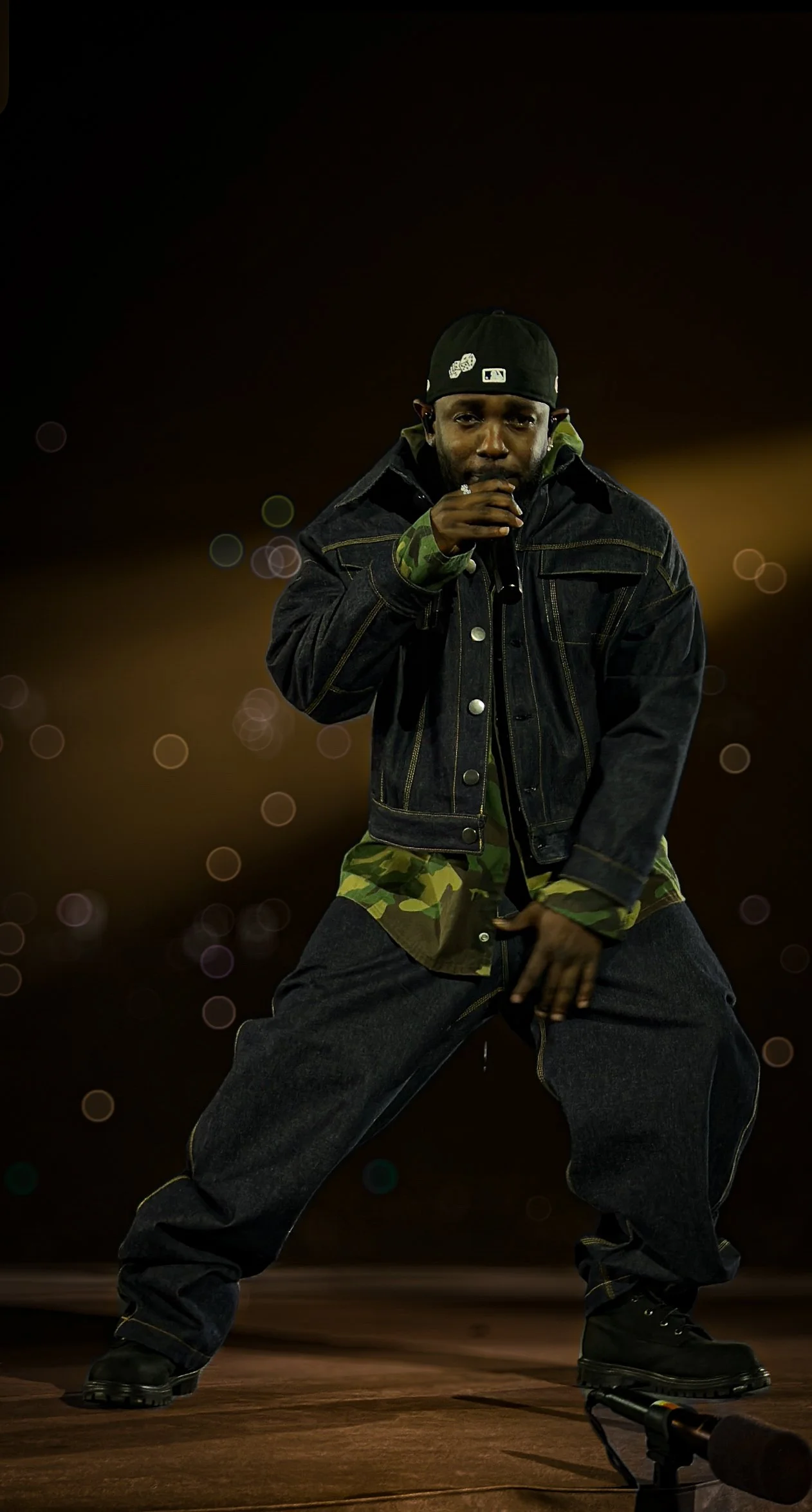 A man performing on stage, holding a microphone, dressed in a black jacket, camouflage shirt, baggy jeans, and black boots, with a dark background and bokeh lights.