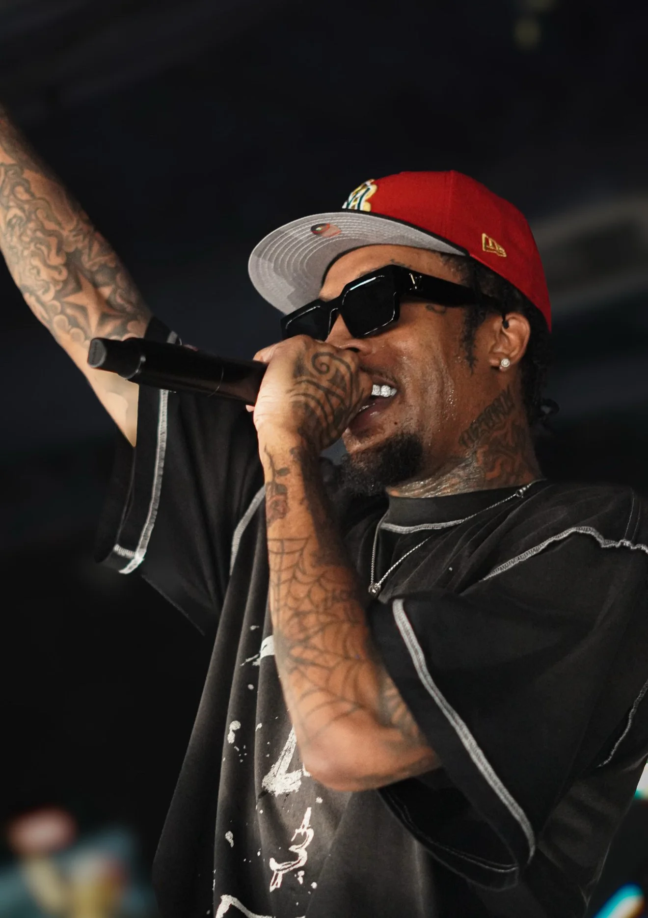 A male rapper with tattoos, sunglasses, and a red and gray baseball cap, performing with a microphone.