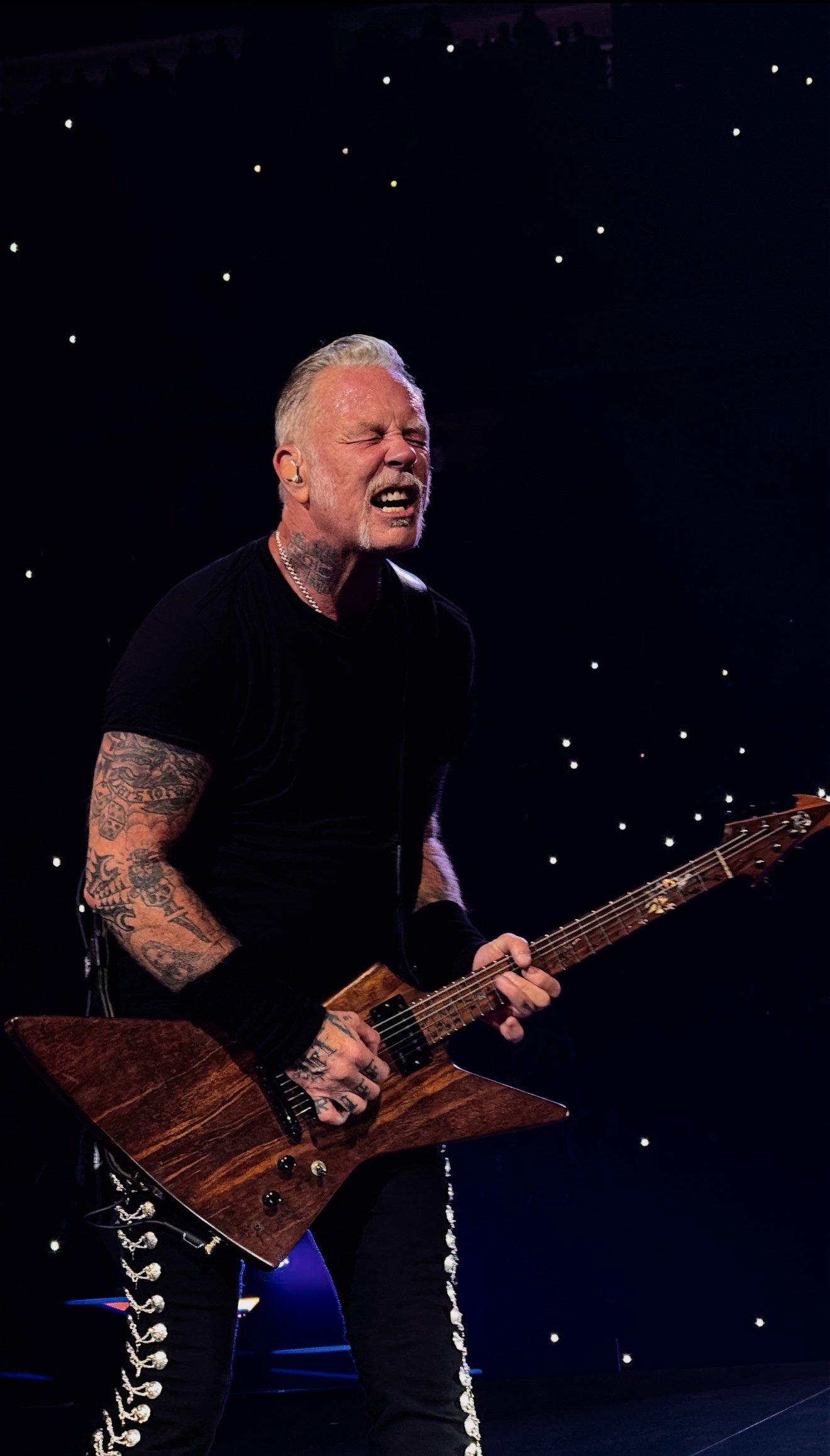 A male guitarist with tattoos, gray hair, and a beard performing passionately on stage with a dark background and small bright lights.