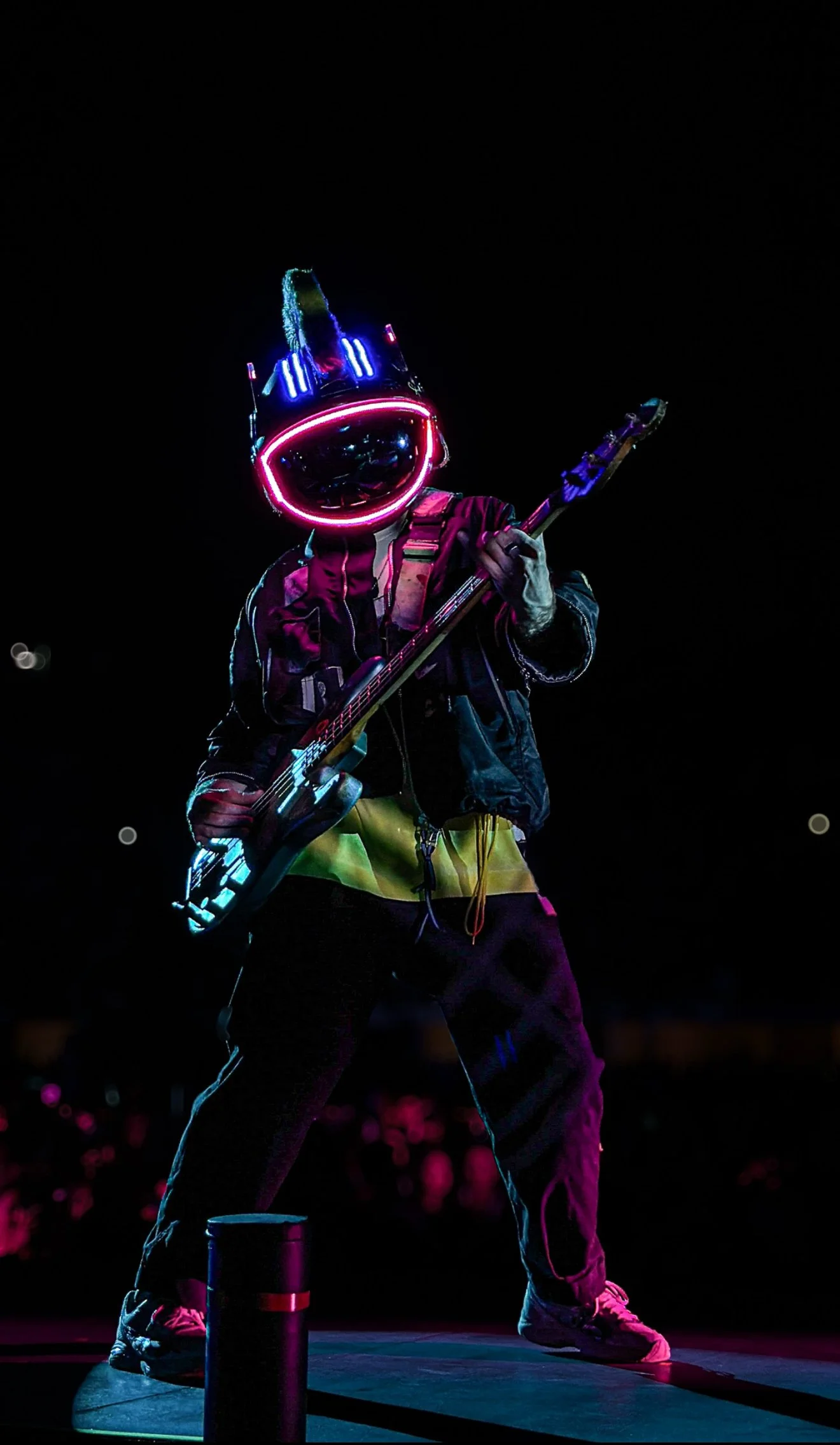 Person wearing a glowing helmet with neon lights, playing an electric guitar on a dark stage.
