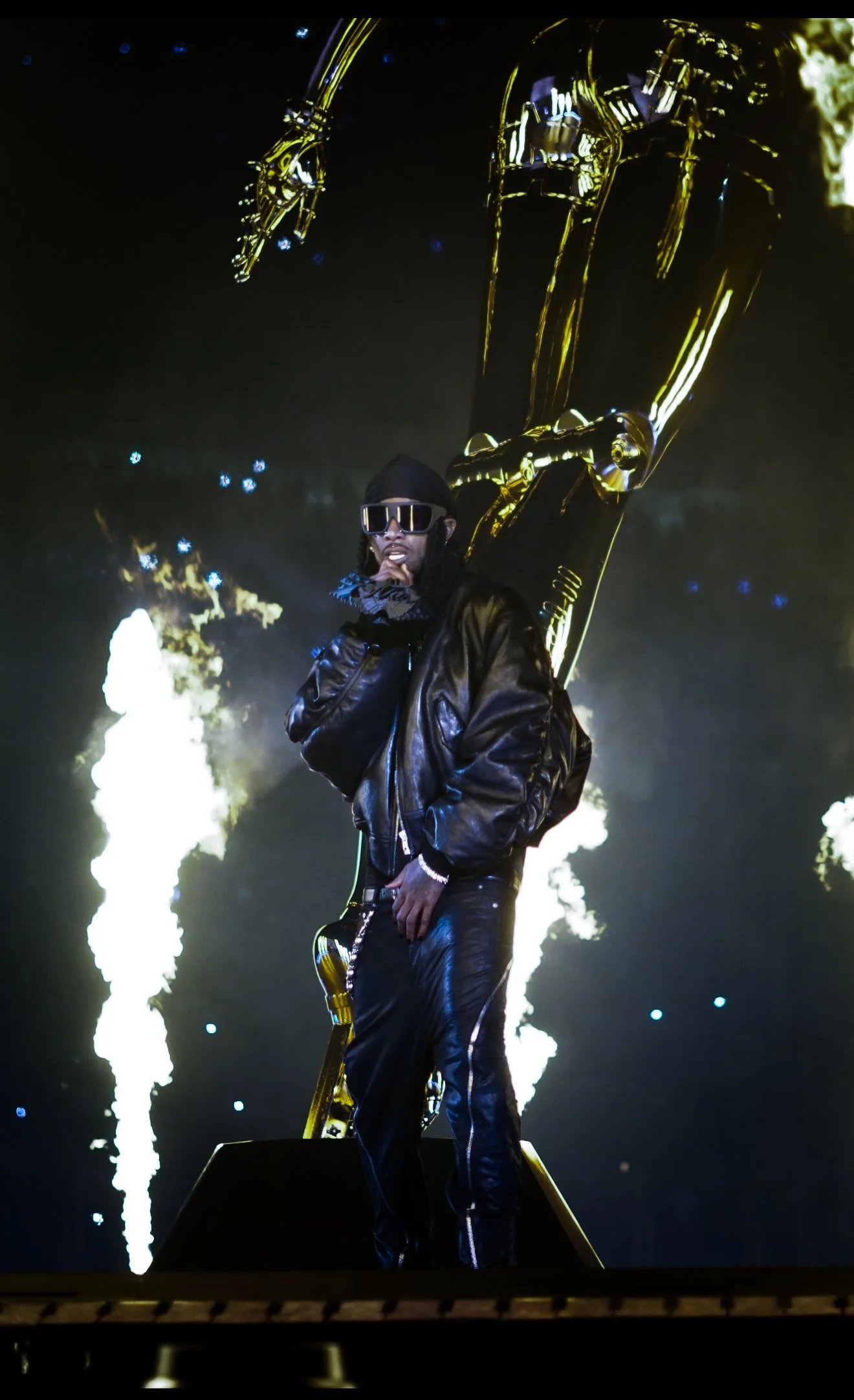 Musician standing on stage with a large golden sculpture behind him, surrounded by colorful lights and smoke effects, wearing black leather outfit and sunglasses.