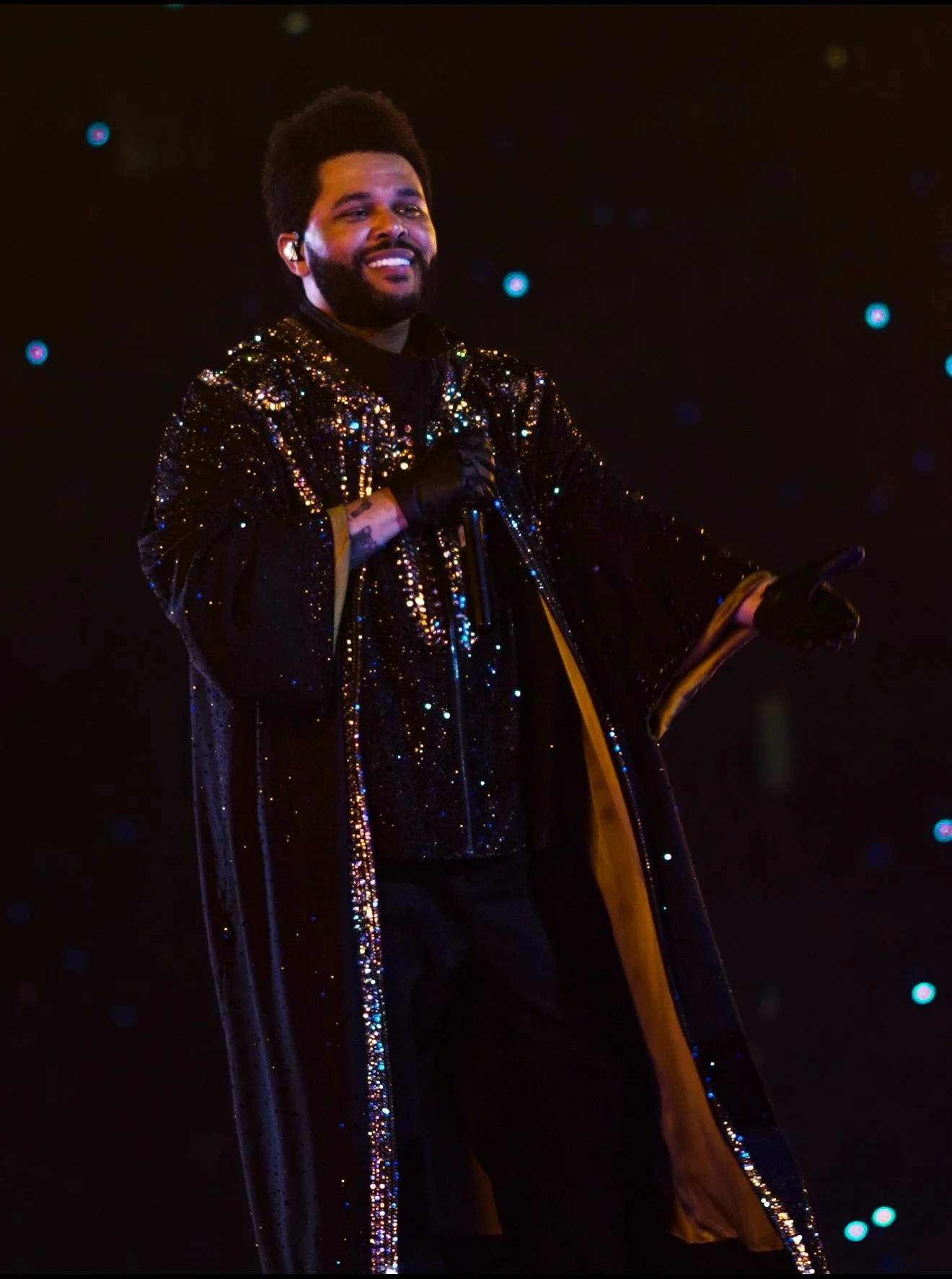 A man with a beard and afro hairstyle smiling while holding a microphone on stage. He is wearing a black, glittery, sequined cape and gloves, with a black outfit underneath. The background is dark with small blue lights scattered.