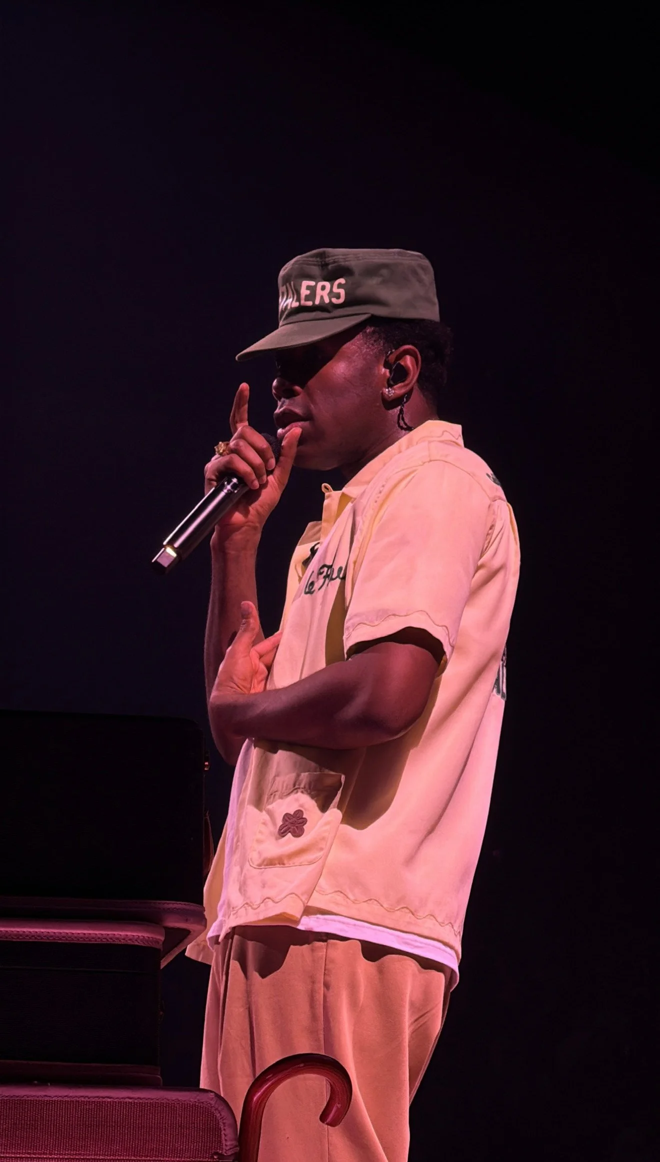 A person on stage holding a microphone, wearing a beige shirt, beige pants, a baseball cap with 'RAILERS' written on it, and sunglasses, with a dark background.