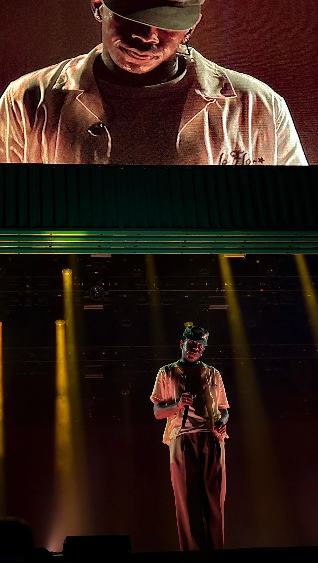 Performer on stage during a concert or show, holding a microphone, under yellow stage lights, with a large screen showing a close-up of their face behind him.