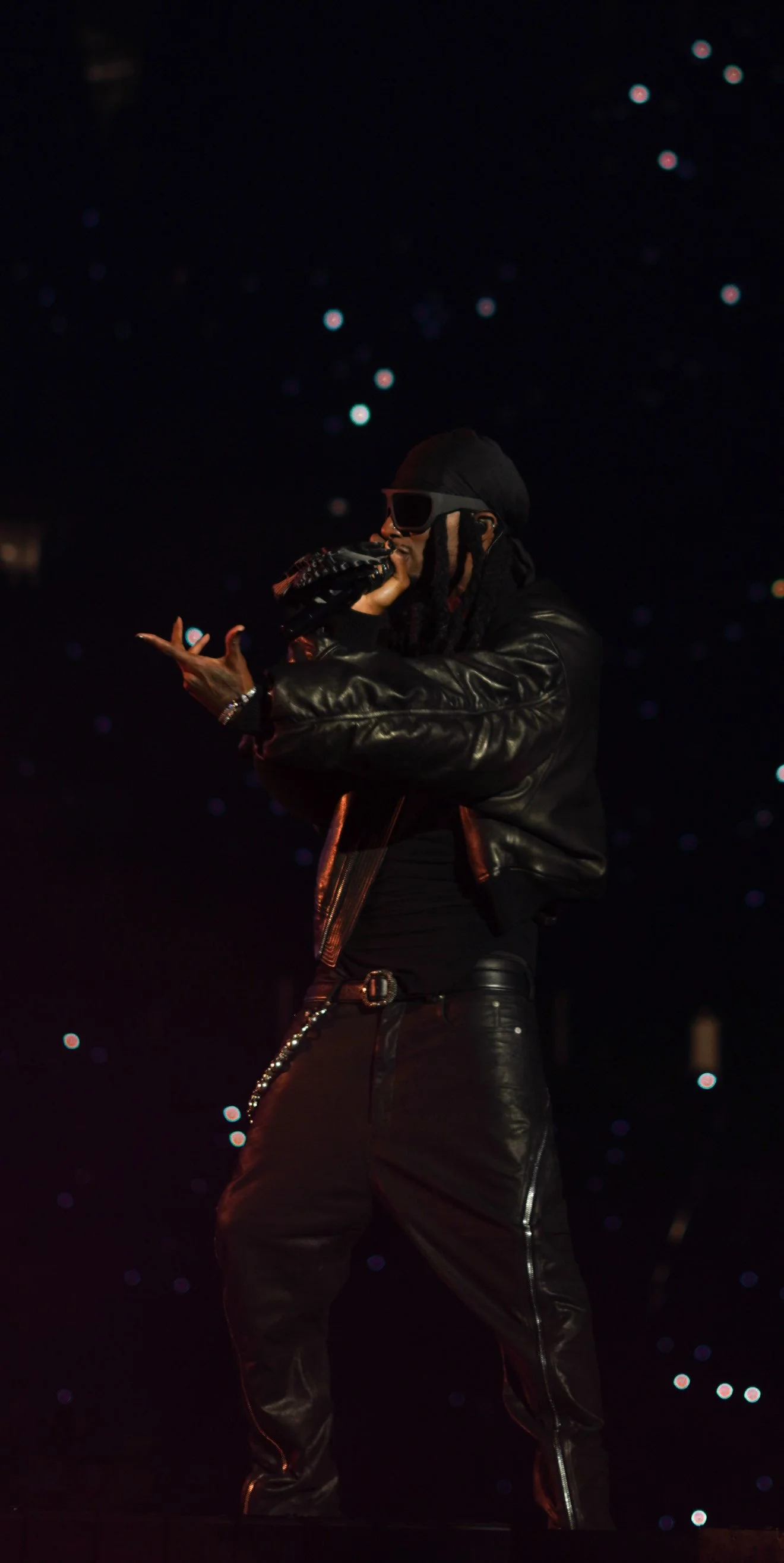 A male performer dressed in black leather clothing, wearing a black head covering and sunglasses, singing into a microphone on stage with a starry background.