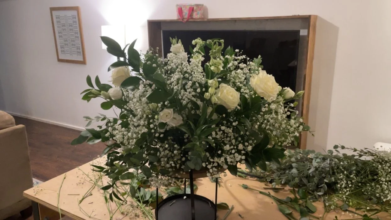 A bouquet of white roses, baby's breath, and greenery on a table, with some floral and greenery pieces scattered around.