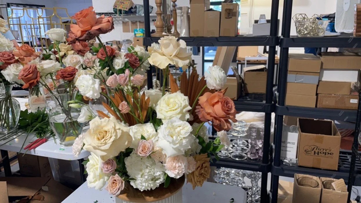 A large floral arrangement with white, blush pink, peach, and cream-colored roses and flowers in a brown vase, set on a white table in a warehouse or floral shop with shelves of boxes, glassware, and supplies around.
