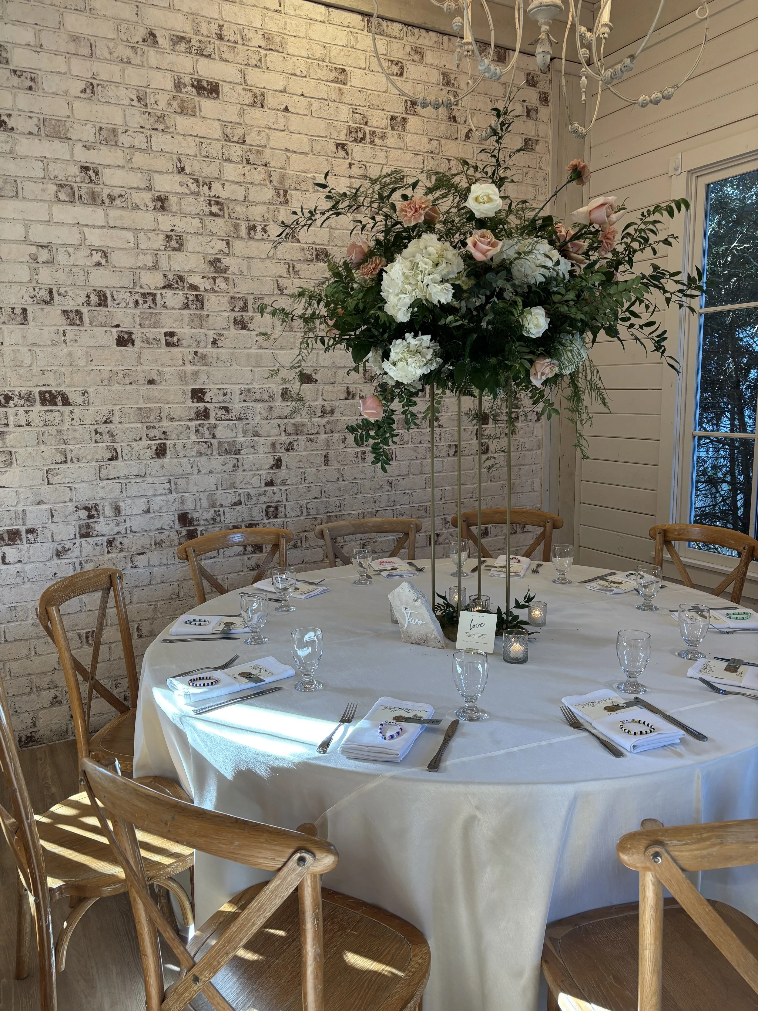 Round table set for a formal event with a tall floral centerpiece, white tablecloth, napkins, silverware, wine glasses, and small candles, with wooden chairs surrounding the table and a brick wall in the background.
