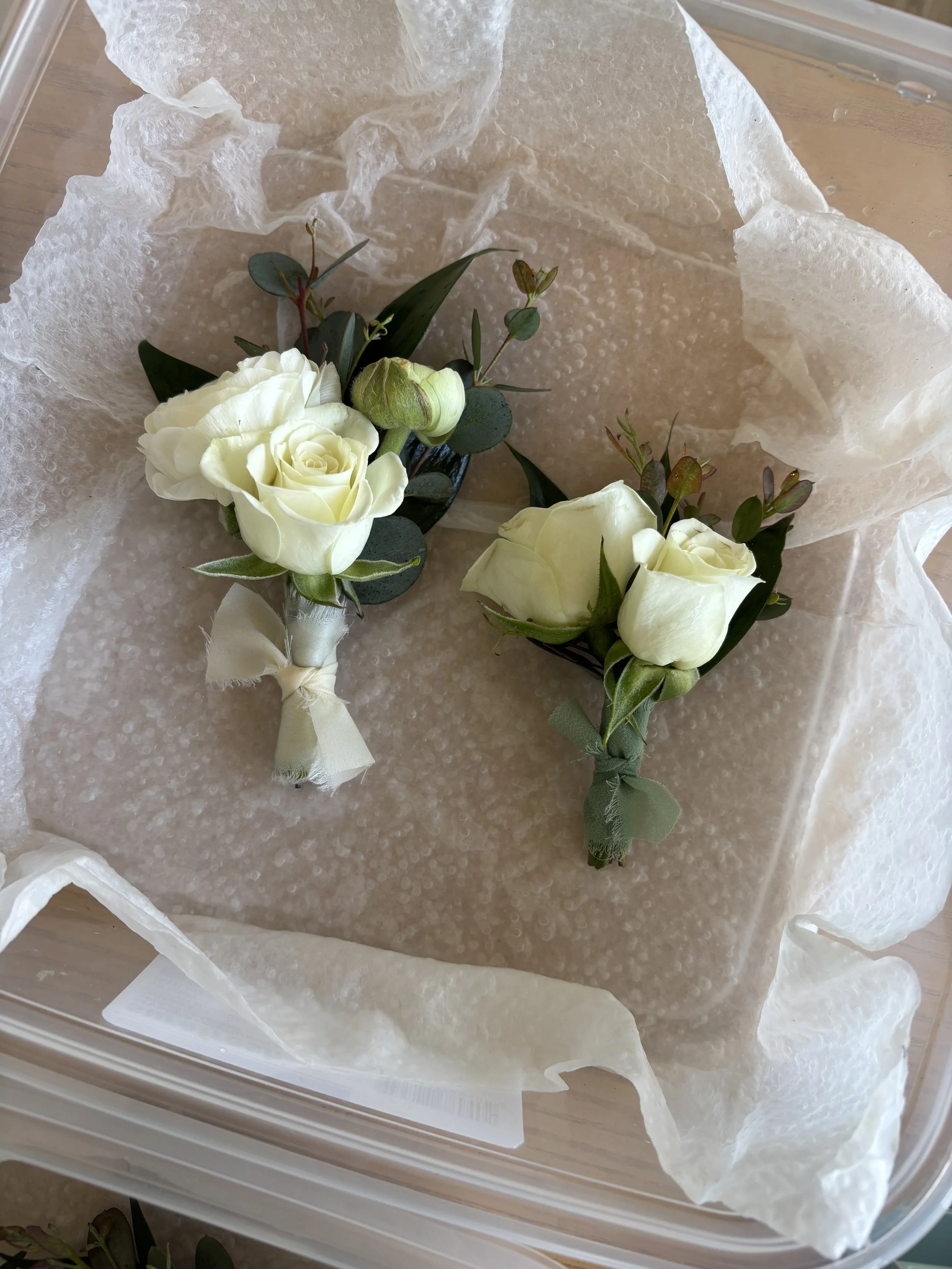 Two white boutonniere floral arrangements with green leaves, wrapped in light-colored ribbon, resting on protective bubble wrap and tissue paper inside a clear plastic box.