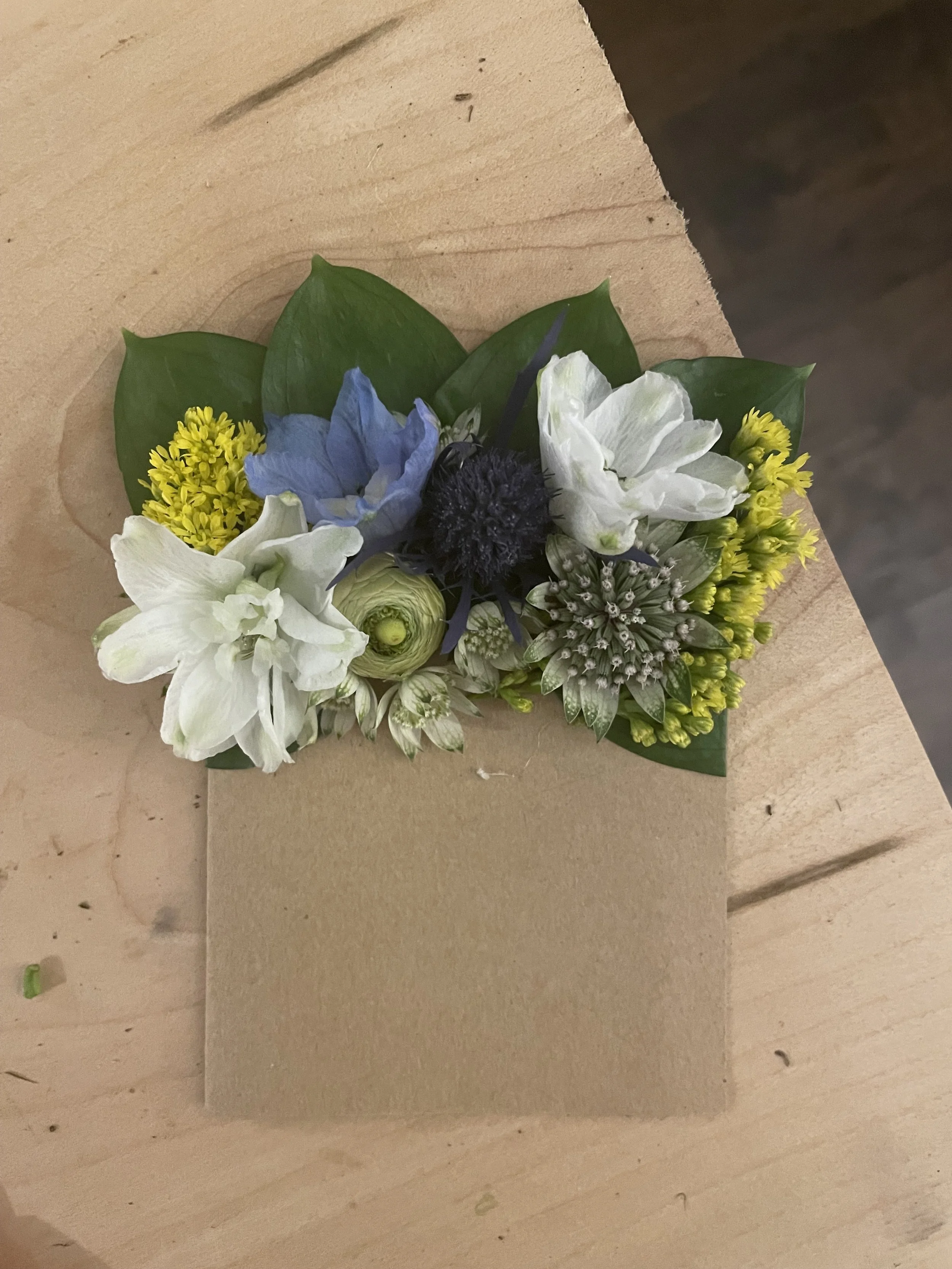 A boutonniere floral arrangement with white, blue, and yellow flowers, green leaves, and a dark central flower, resting on a cardboard box on a wooden surface.