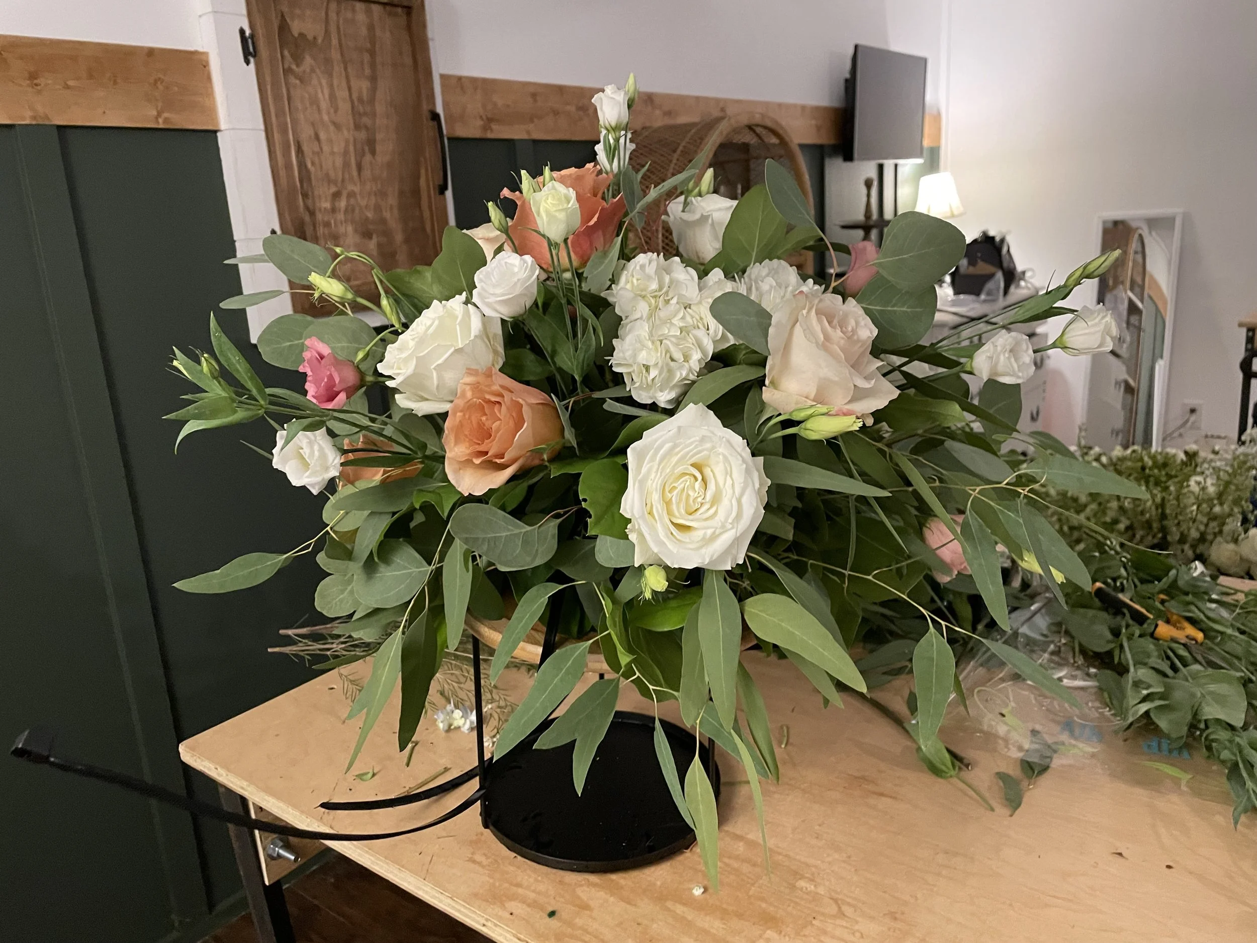 A floral arrangement with white, peach, and pink roses, white lisianthus, and greenery on a black stand, placed on a wooden table indoors.