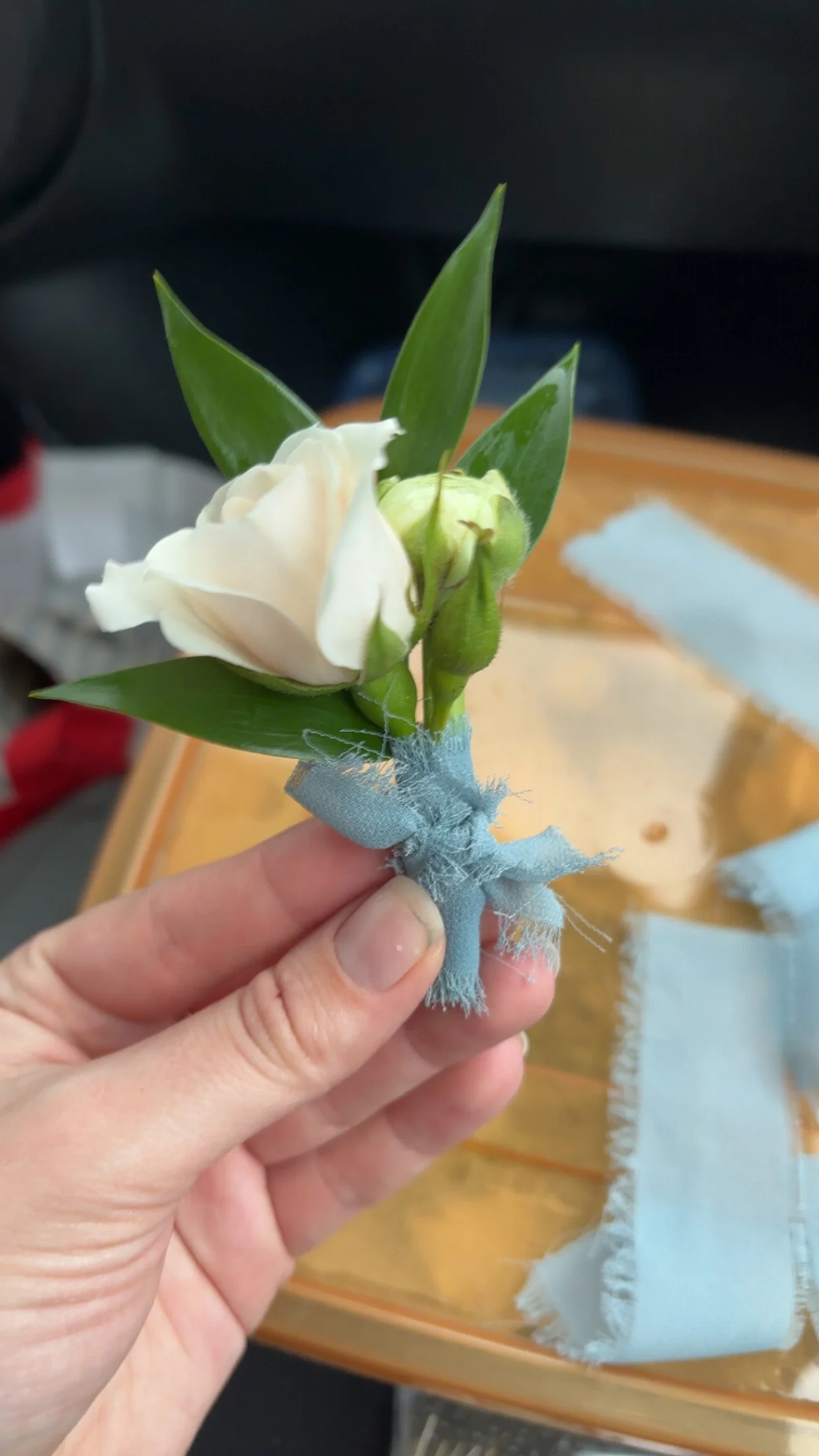 Hand holding a small boutonniere with a white flower and green leaves, wrapped with blue fabric.