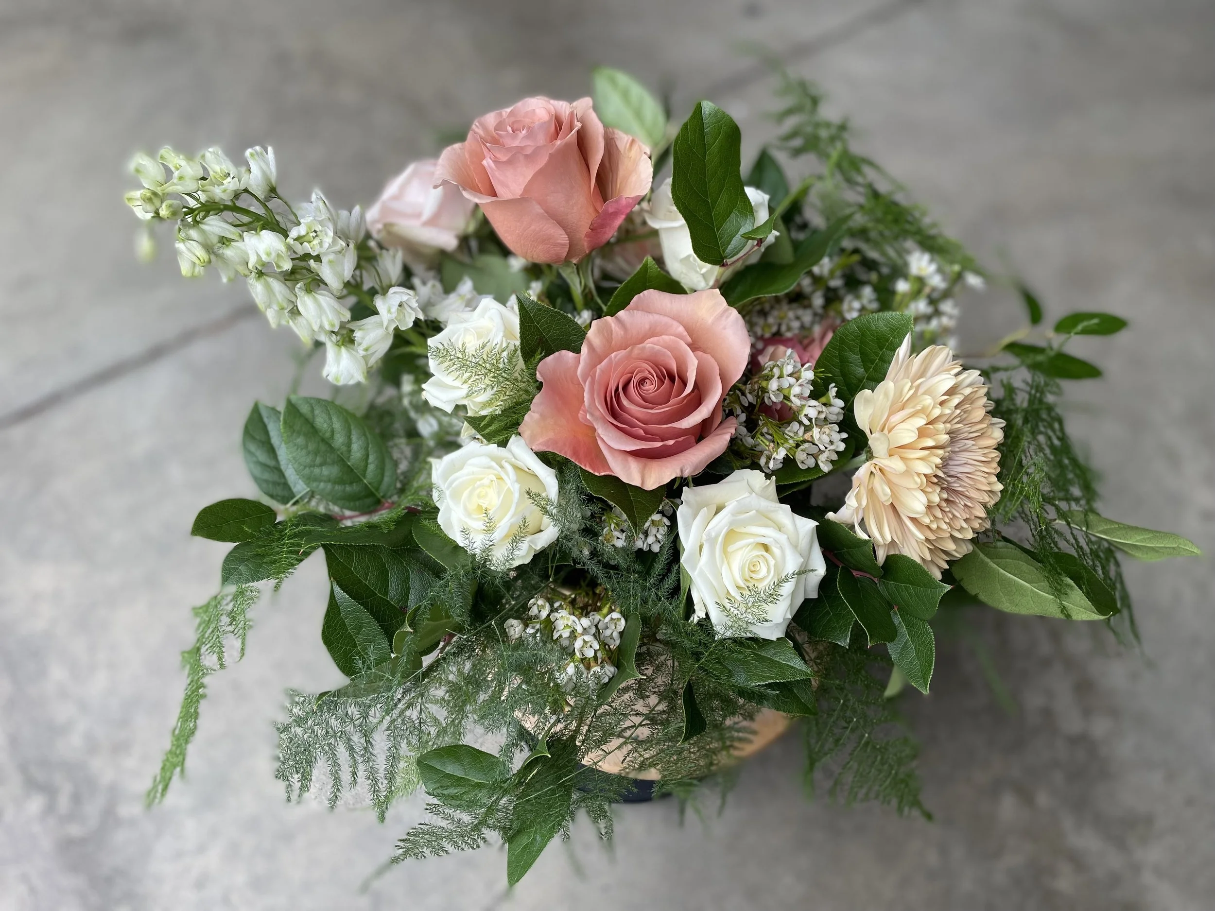 A bouquet of pink, white, and peach roses with white daisies, greenery, and small white flowers, arranged in a wicker basket on a concrete surface.