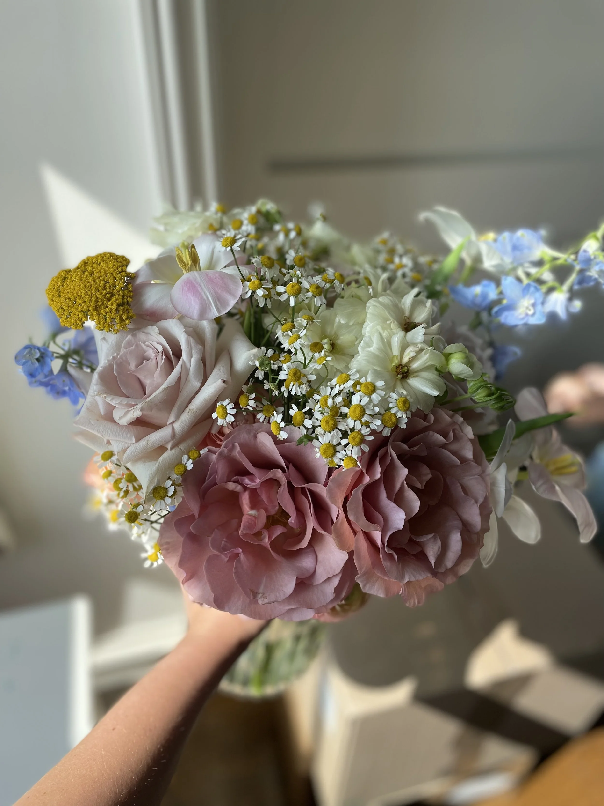 Hand holding a bouquet of pink, white, and blue flowers near a window with sunlight.