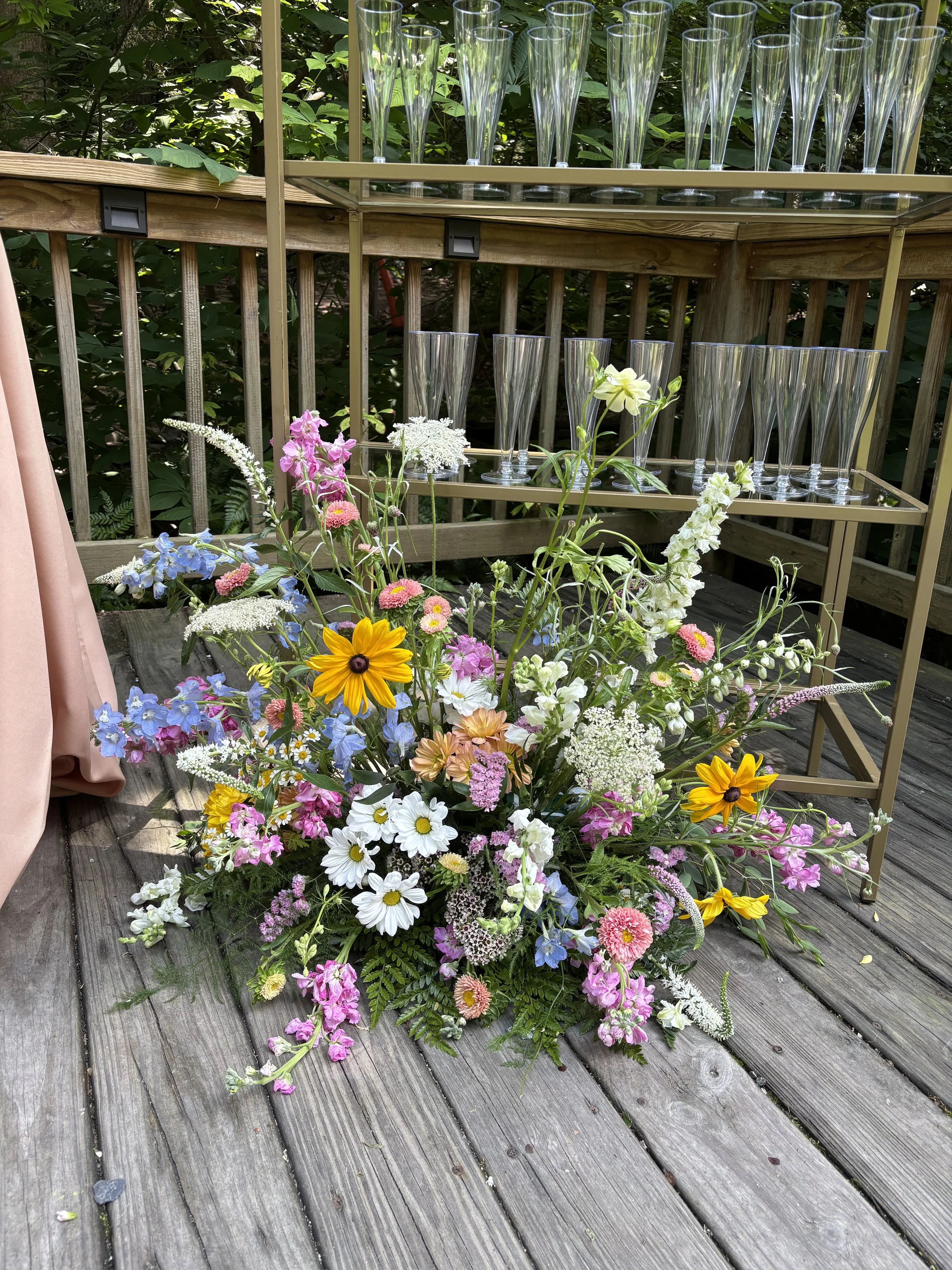 Colorful floral arrangement in pink, yellow, white, purple, and blue flowers on a wooden deck with glassware on a two-tiered wooden stand against a green leafy background.
