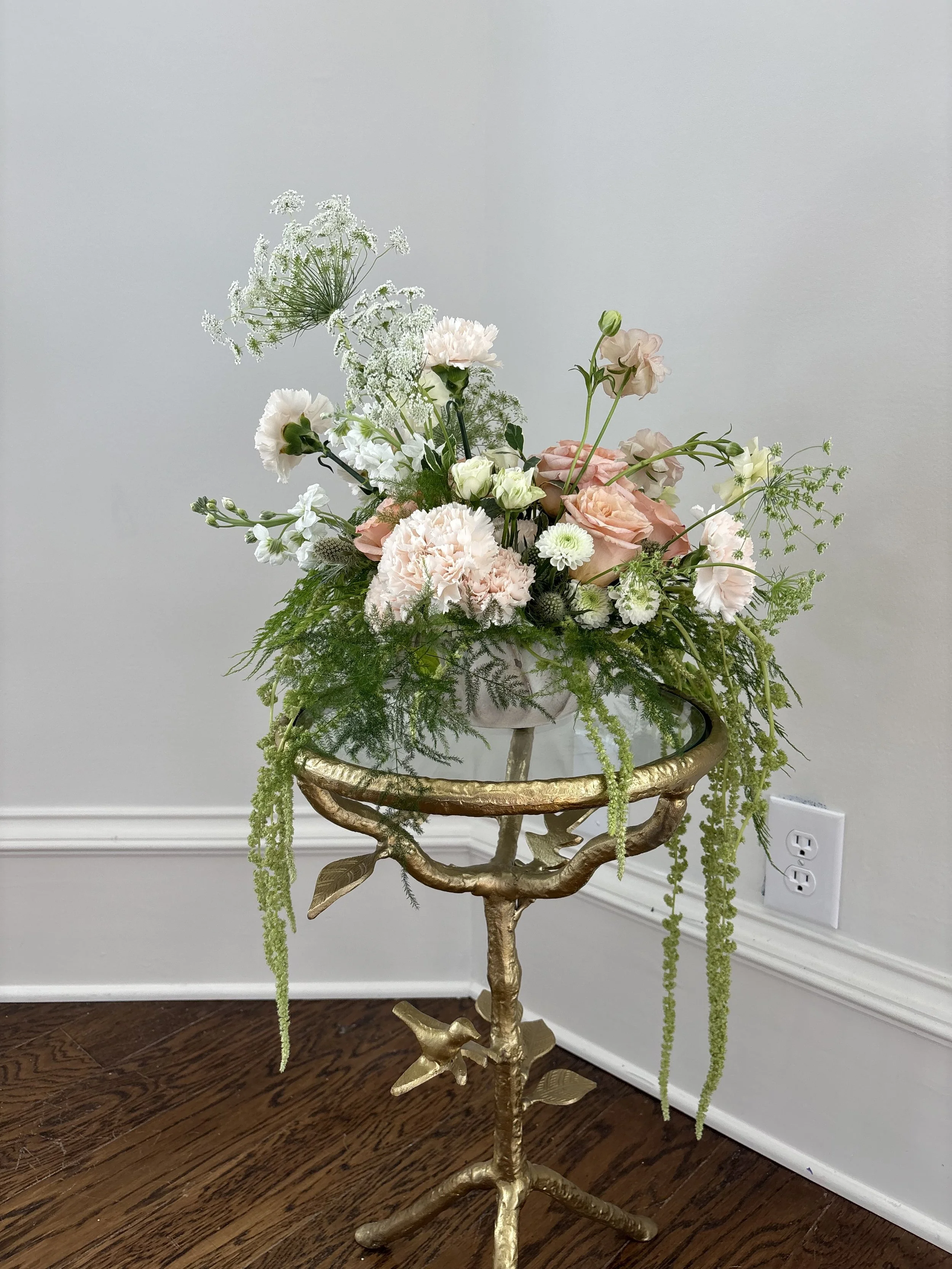 A gold ornate plant stand with a floral arrangement of pink, white, and cream-colored flowers and green foliage on a wood floor next to a white wall with electrical outlets.