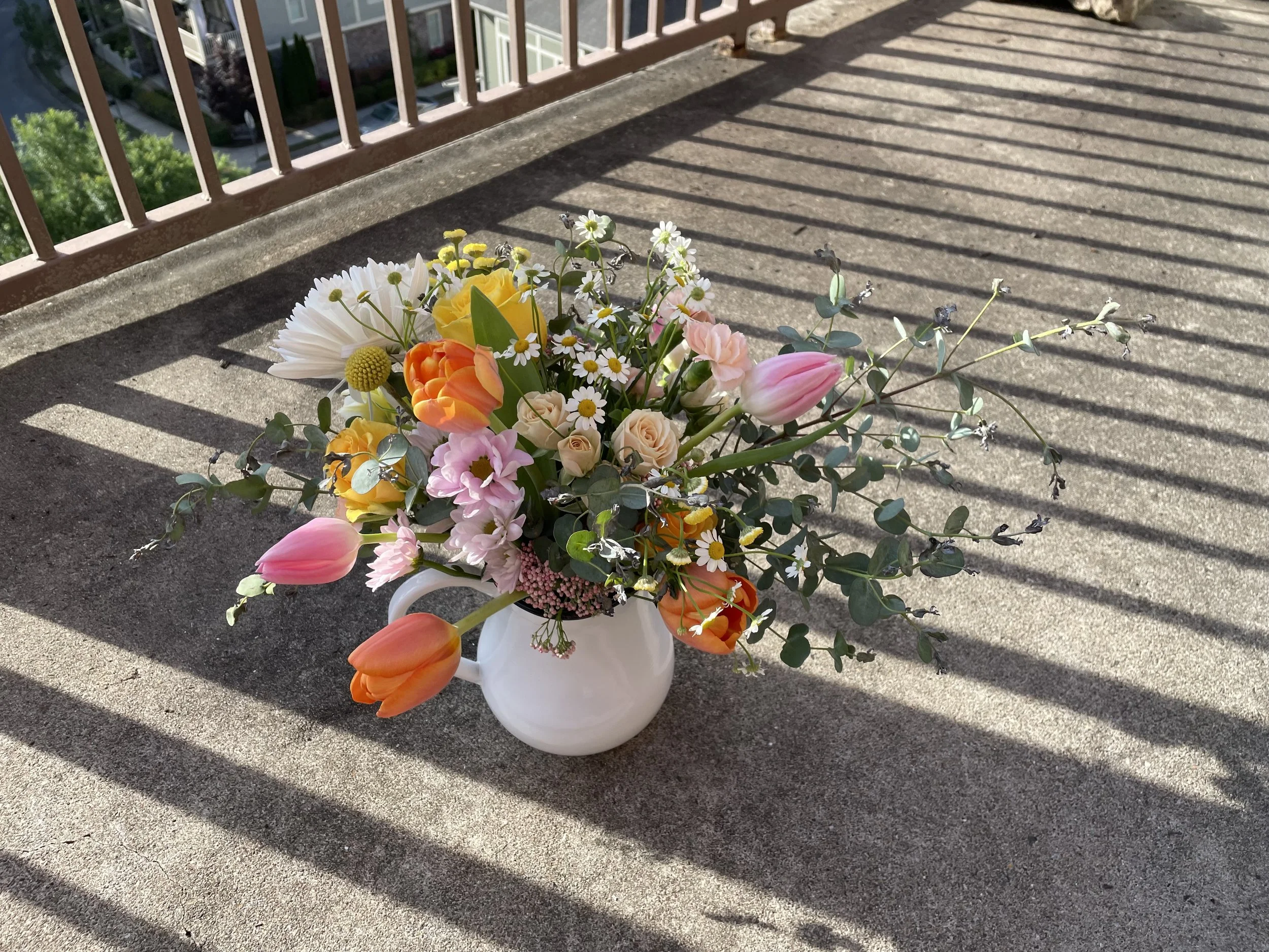 A white vase with a colorful assortment of flowers, including tulips, daisies, roses, and greenery, sitting on a concrete balcony with shadows cast by a railing.