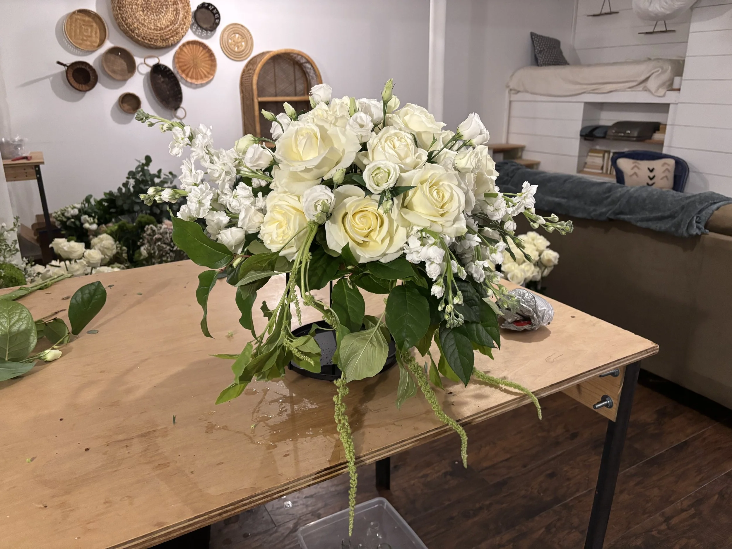 White floral arrangement with roses and other white flowers on a wooden table in a cozy room with wall decorations and a sofa.