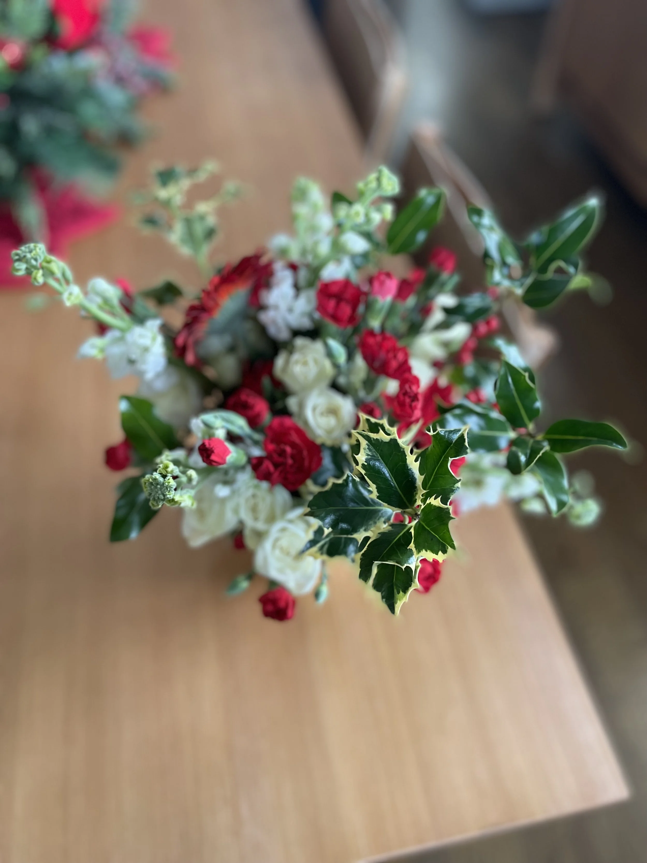 Holiday floral arrangement with white and red flowers, green holly leaves with yellow edges, and other greenery on a wooden table.