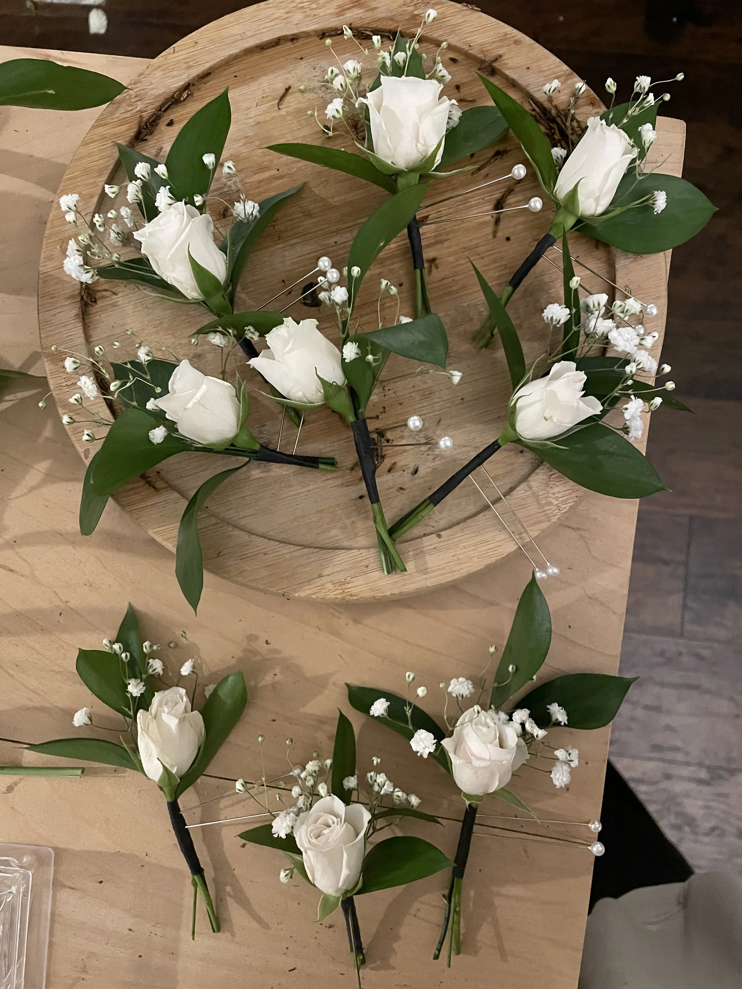 Bouquet of white roses, baby's breath, and green leaves arranged with black pins on a round wooden board.