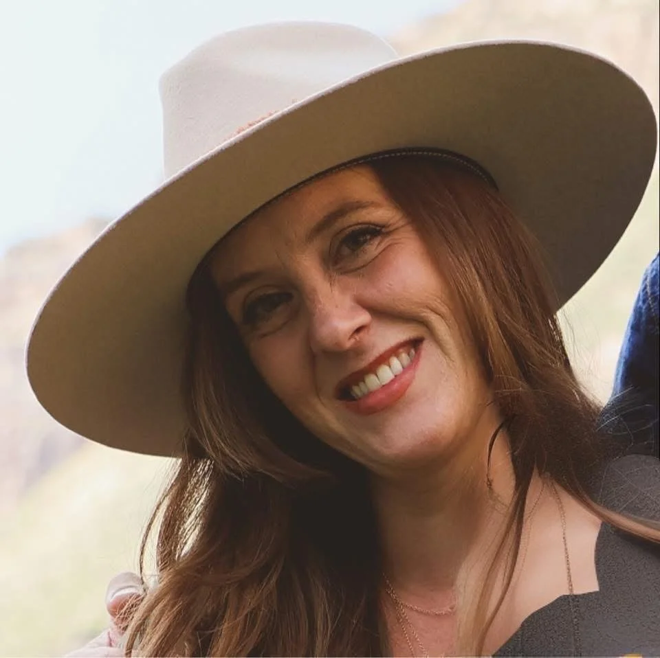 A woman with long auburn hair smiling, wearing a large beige hat and black clothing outdoors.