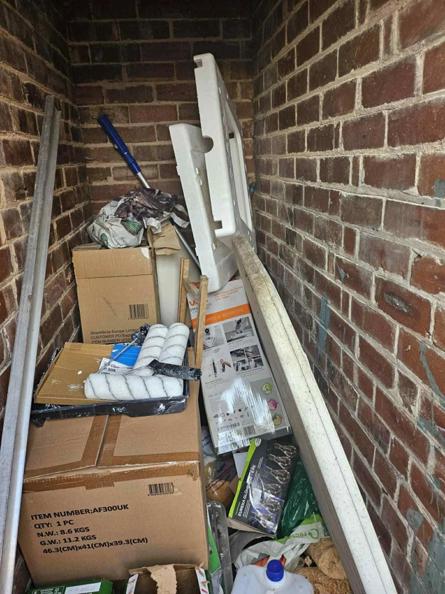 A cluttered storage space with boxes, paint rollers, a ladder, and various tools and supplies, all packed into a narrow brick-walled storage area.