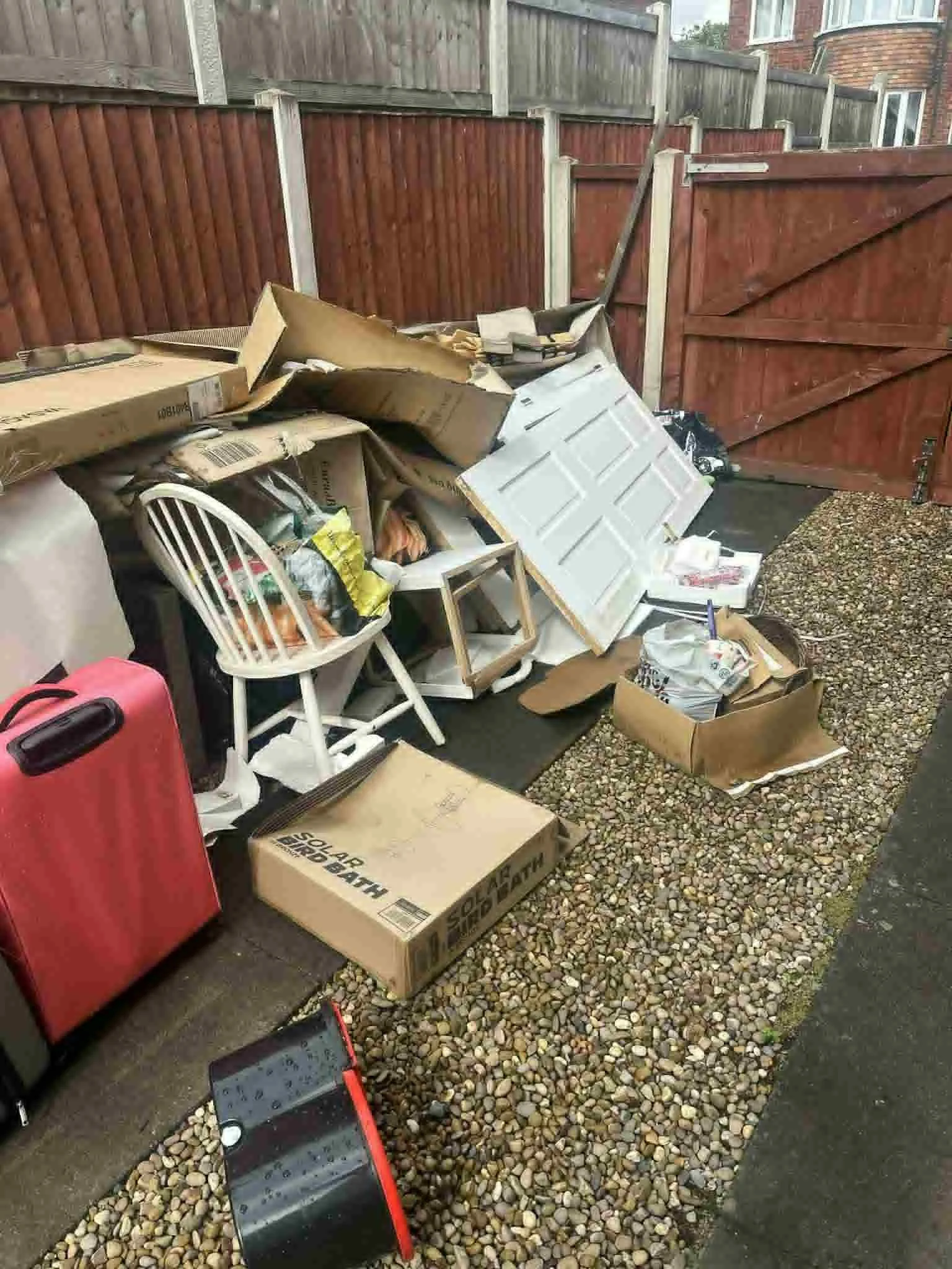 Yard filled with discarded boxes, furniture, and household items behind a wooden fence door.