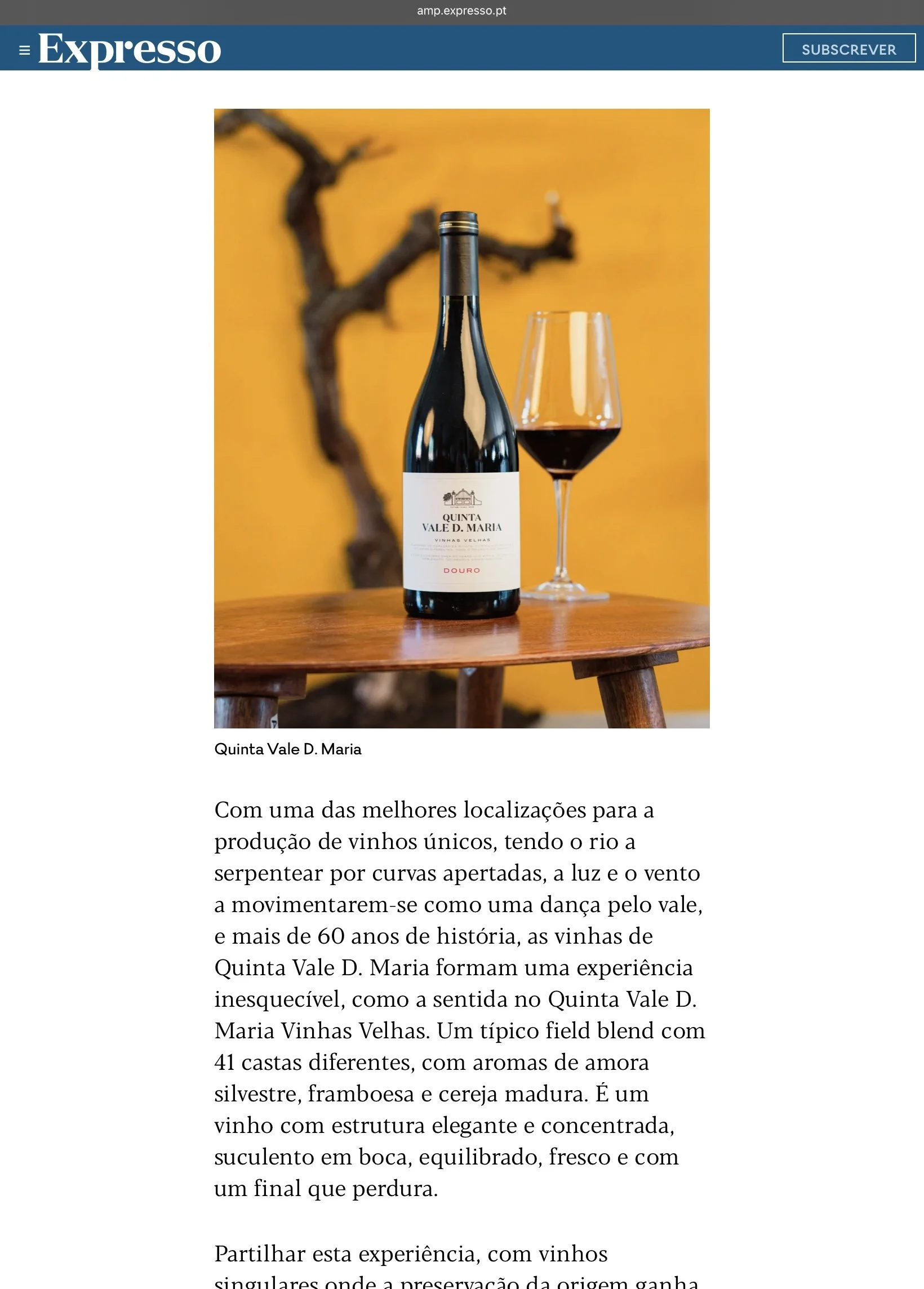 Fotografia de Matheus Suliman aplicada na imprensa. Garrafa de vinho tinto Quinta Vale D. Maria e copo de vinho em uma mesa de madeira, com fundo amarelo e videira seca ao fundo.
