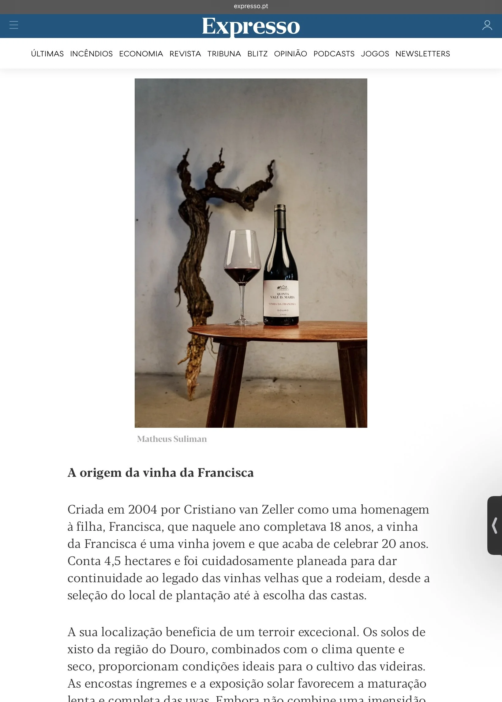 Fotografia de Matheus Suliman aplicada na imprensa. Copo de vinho tinto, garrafa de vinho Quinta Vale D. Maria, mesa de madeira, parede neutra ao fundo, videira seca ao fundo.