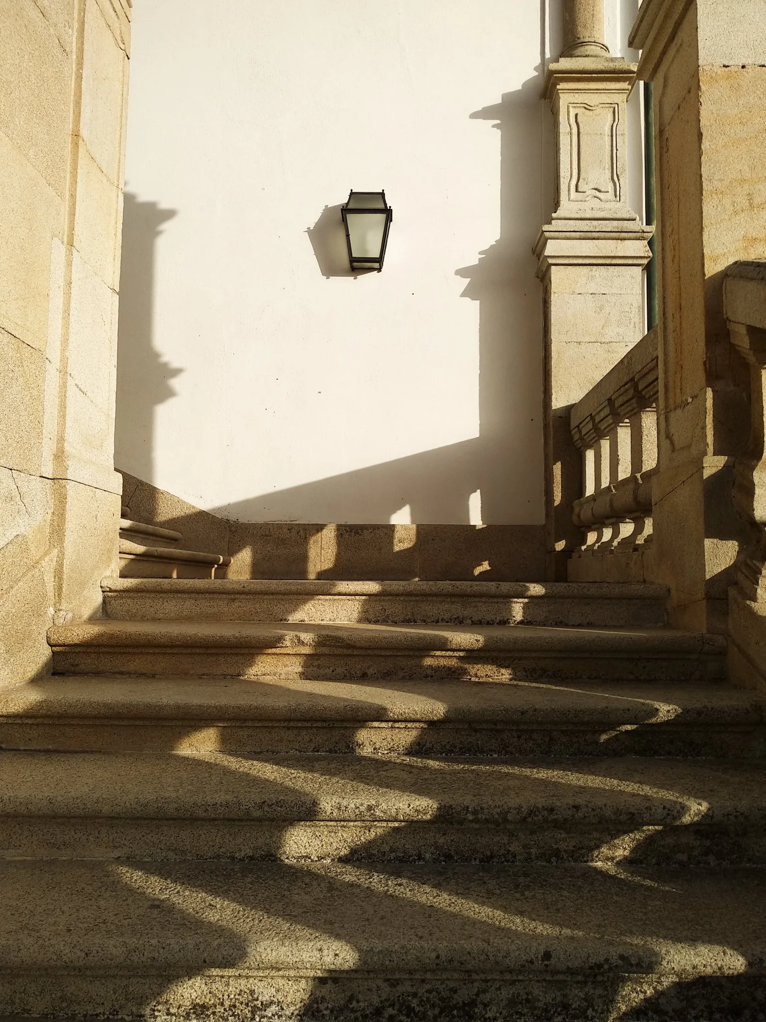 Escadaria de pedra com sombras, parede branca ao fundo, luminária na parede e varanda com balaustrada de pedra. Fotografia de Viagem por Matheus Suliman