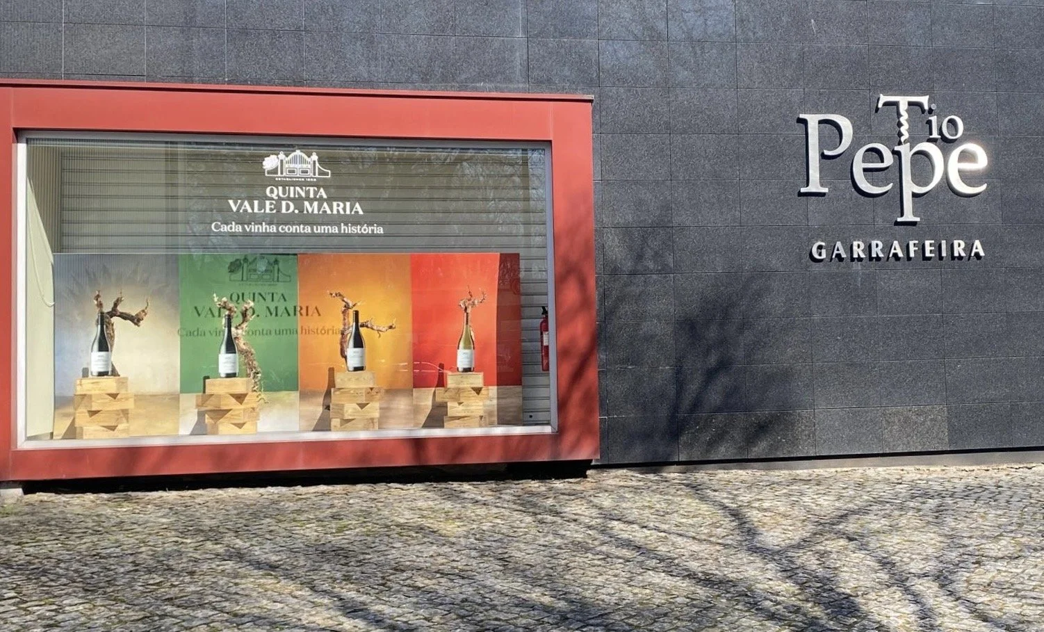 Vitrine mostrando garrafas de vinho em frente às imagens de campanha oficial dos vinhos Quinta Vale D. Maria. As fotos contém videiras, com fundo branco rústico, verde, amarelo e vermelho, em frente a uma loja chamada Tio Pepe Garrafeira no Porto.
