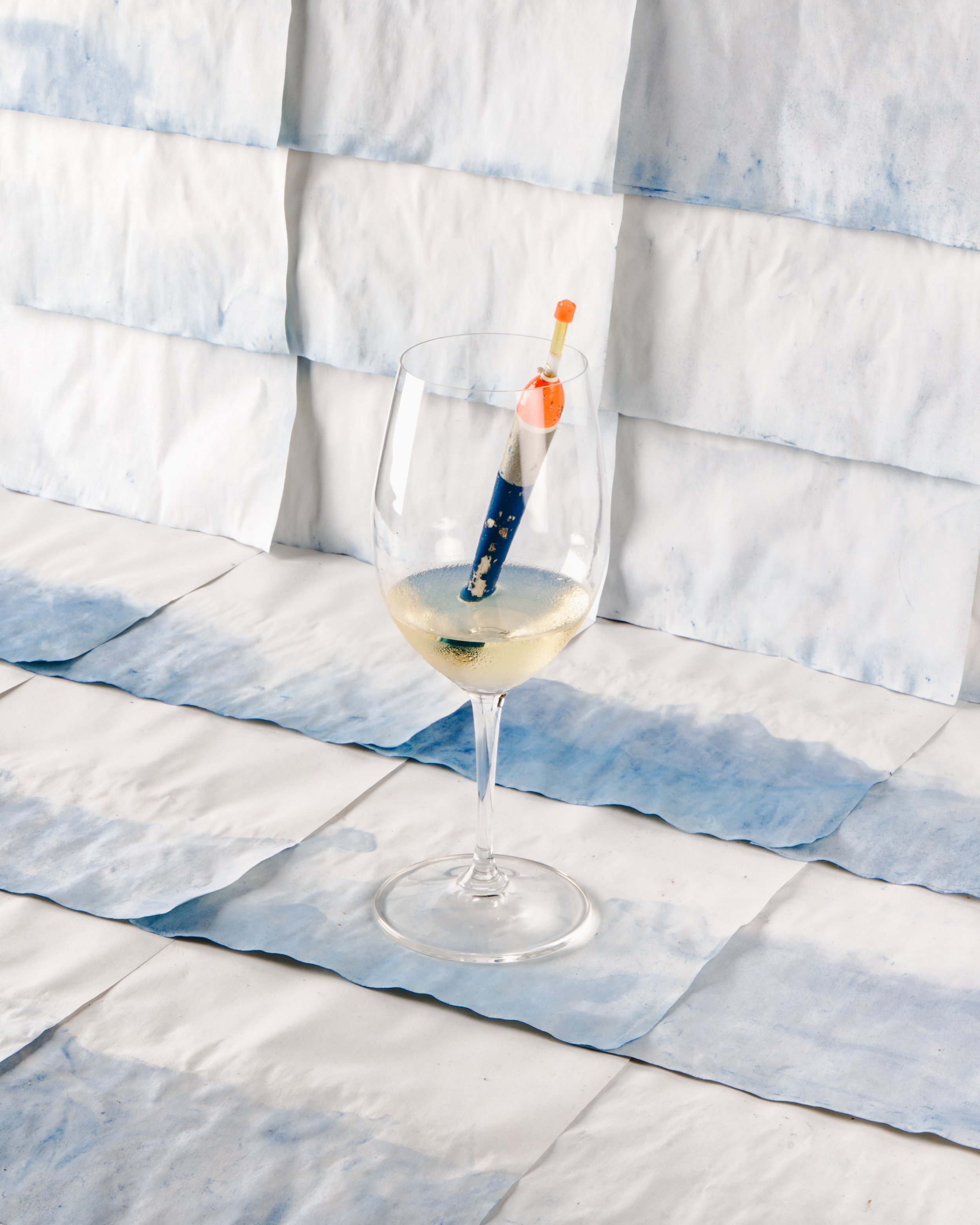 Taça de vinho branco com elemento de pesca dentro do copo, fundo com papeis pintados inspirados nas ondas do mar, fotografia still life por Matheus Suliman