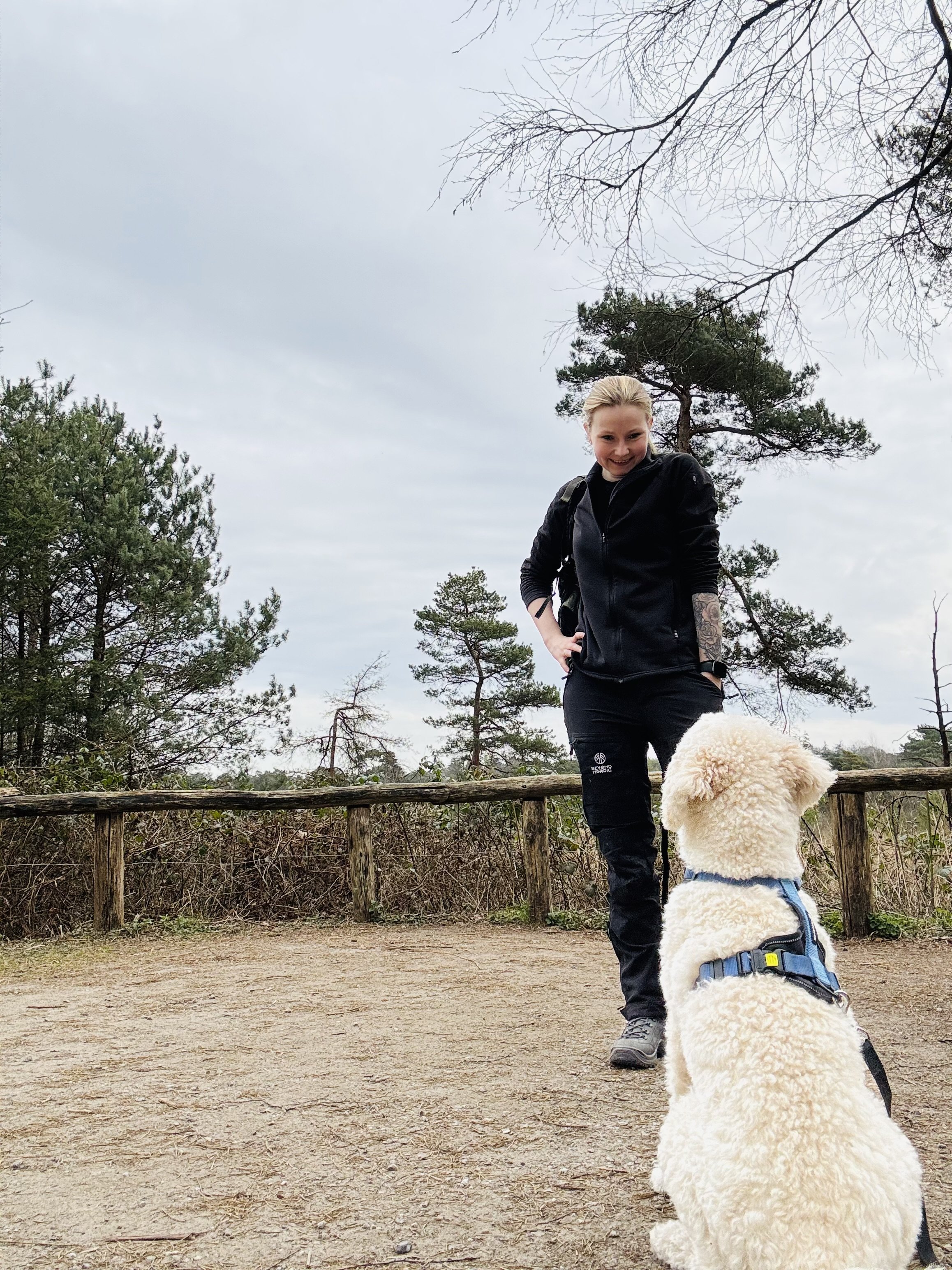 A woman dressed in black hiking attire standing outdoors on a dirt trail, smiling at a seated fluffy white dog wearing a harness, with trees and overcast sky in the background.